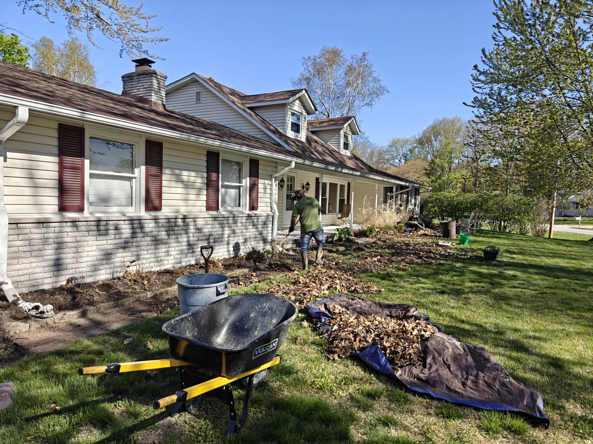 People gardening outside a house on a sunny day, with a wheelbarrow filled with leaves, a tarp with collected leaves, and gardening tools.