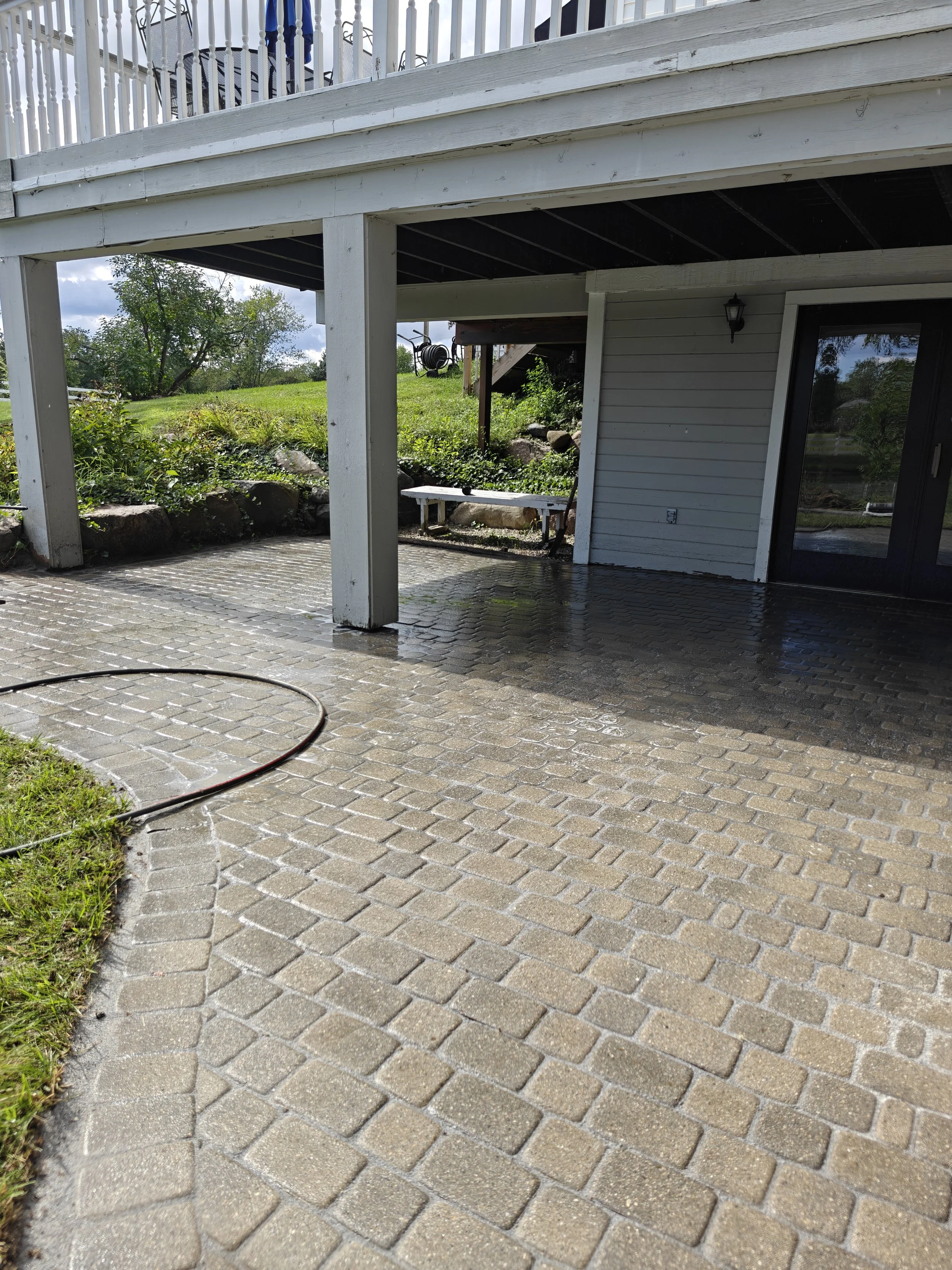 Wet brick patio under a raised balcony with white railing, beside a house with white siding and sliding glass door, outdoor garden with grass, rocks, and tree in the background.