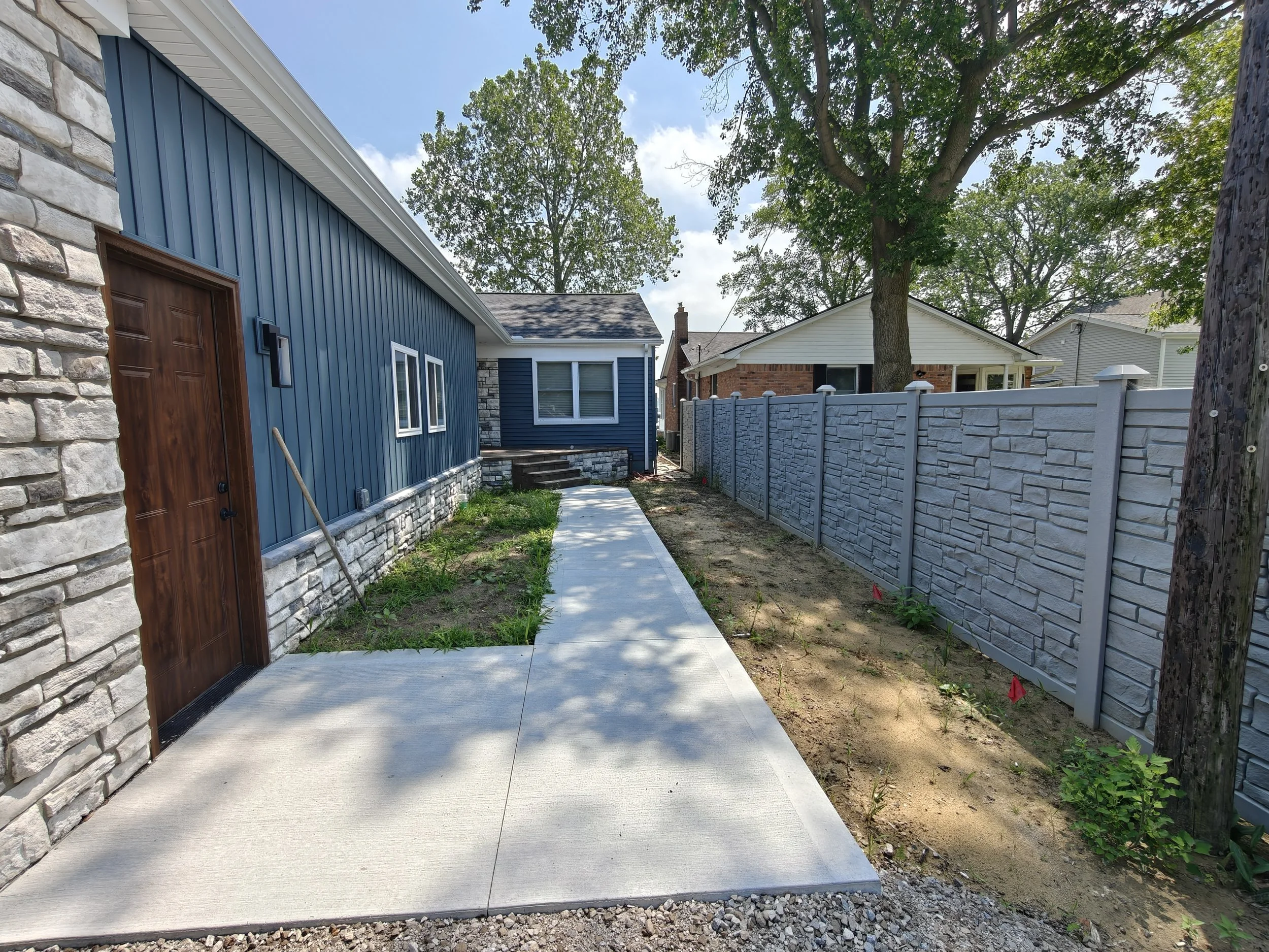 A newly paved concrete sidewalk runs alongside a house with blue siding and stone accents. A wooden door is on the house's side, and a window directly above it. A landscaped area with grass and plants is next to the sidewalk. A tall gray fenced yard 