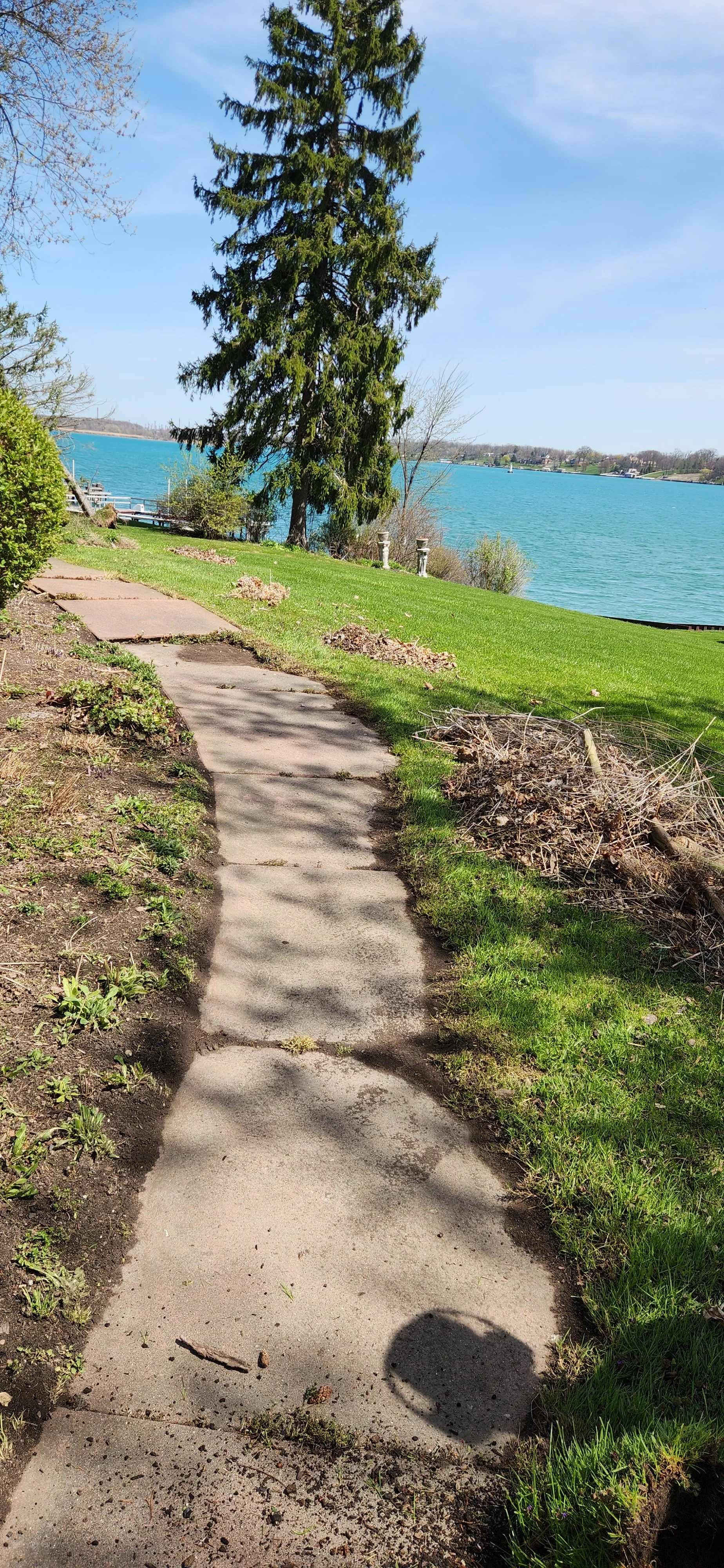 A concrete sidewalk curves through grass and dirt in a backyard, overlooking a large body of water with trees and a clear blue sky in the background.