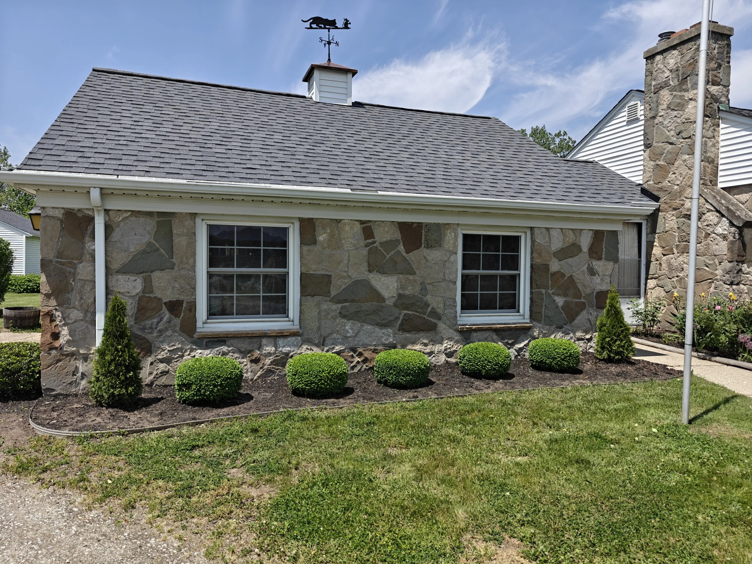 A stone house with two windows, a gray shingle roof with a weather vane of a dog, and a chimney. The front yard has neatly trimmed bushes and small trees, a sidewalk, and a grassy lawn under a partly cloudy sky.