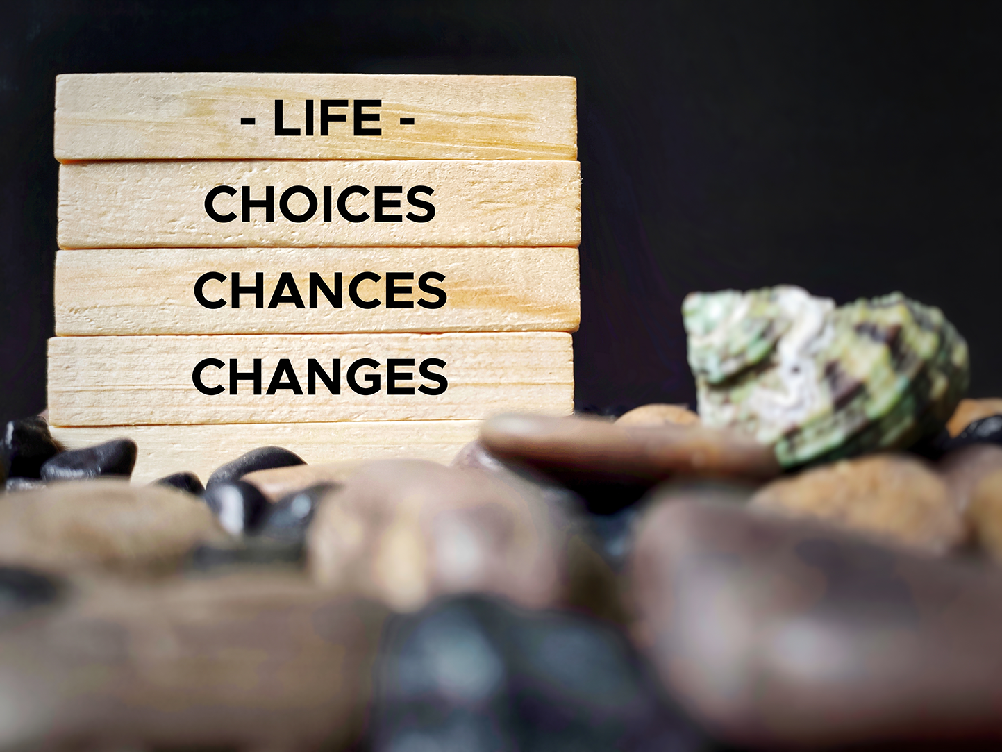 A wooden block display with the words "Life Choices Chances Changes" stacked vertically, surrounded by rocks and a piece of patterned rock, against a black background.