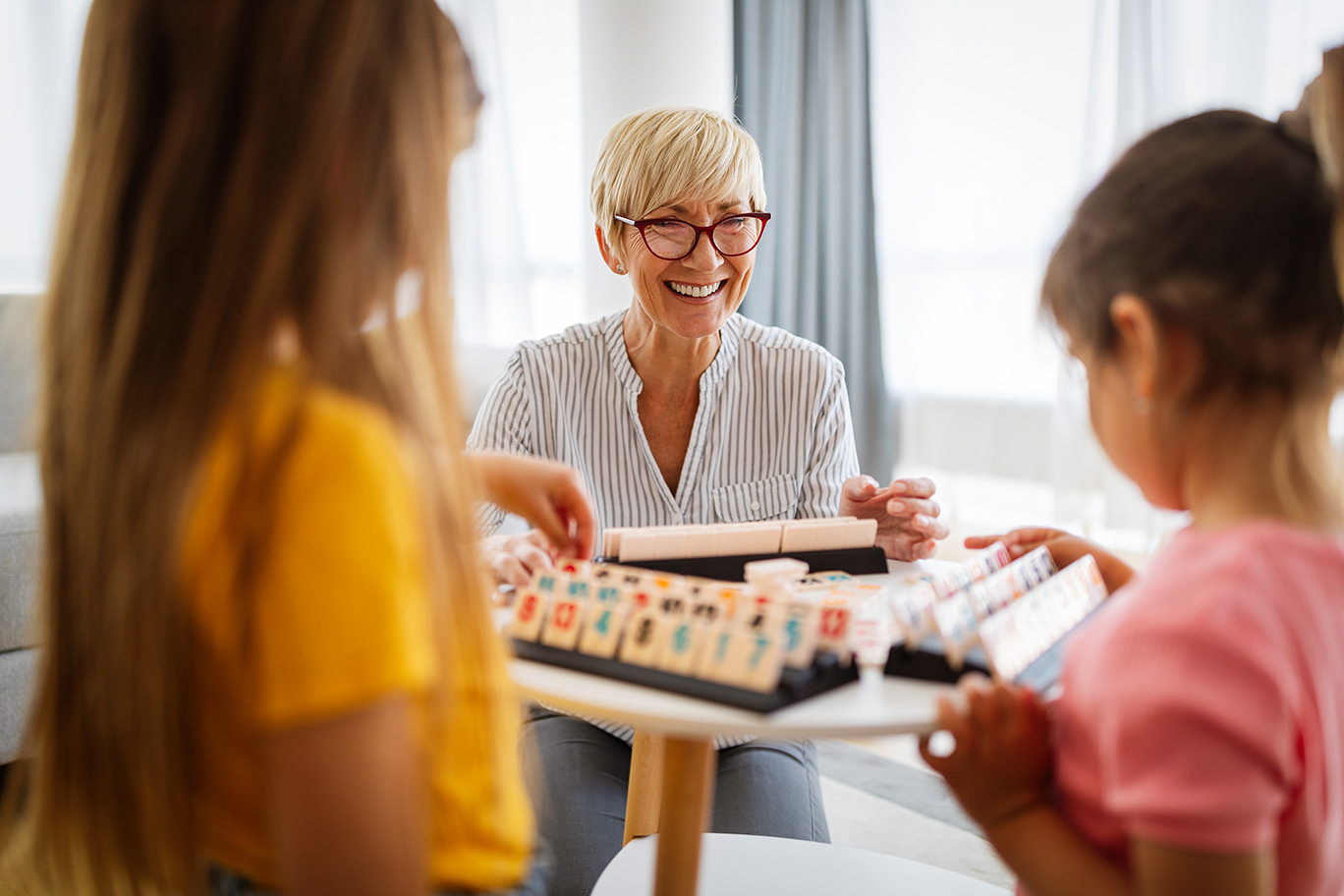 A woman with short blonde hair and glasses smiling and playing a board game with two young girls in a bright living room.