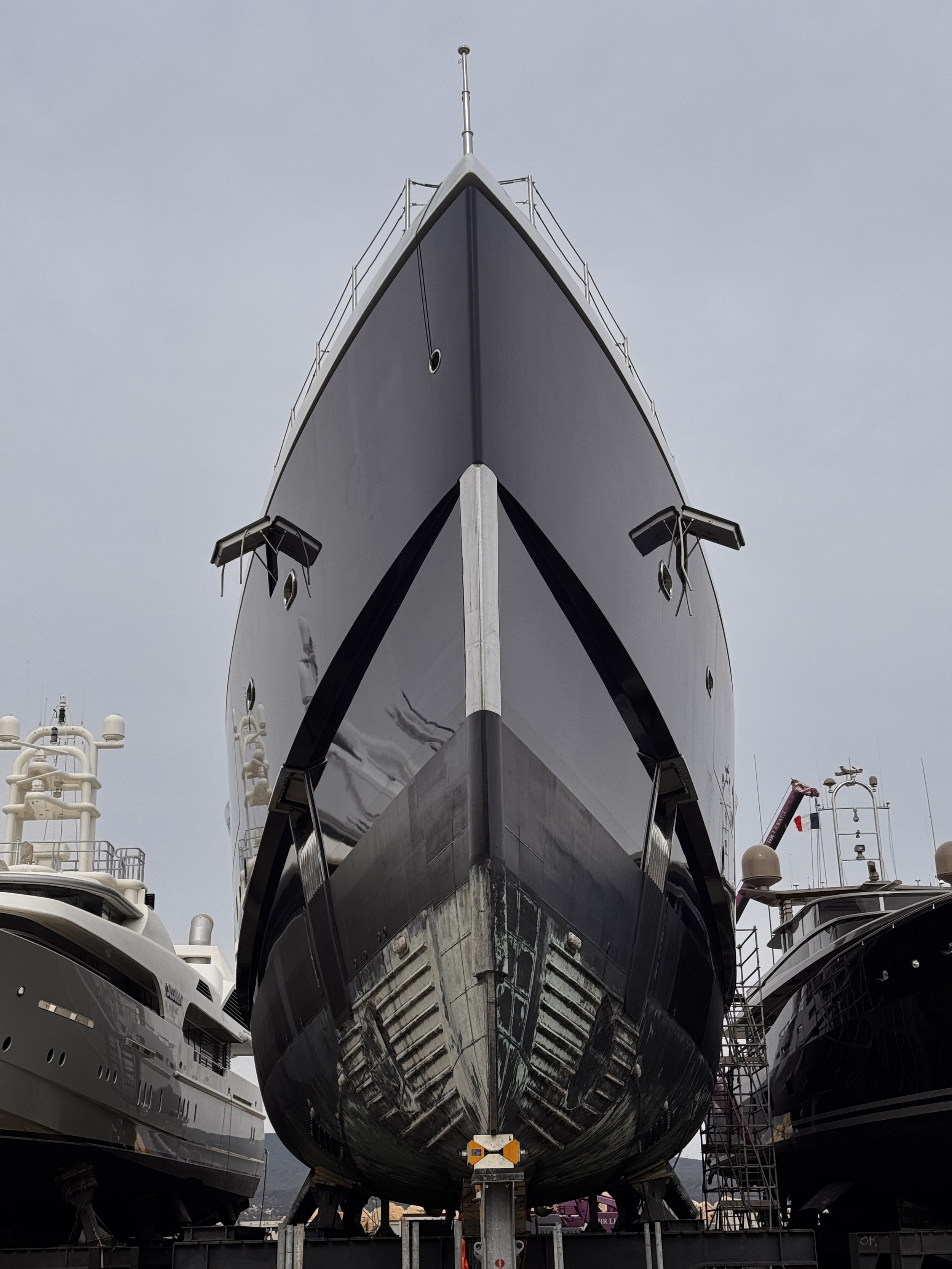Front view of a large yacht on land, flanked by other yachts, with maintenance equipment visible.