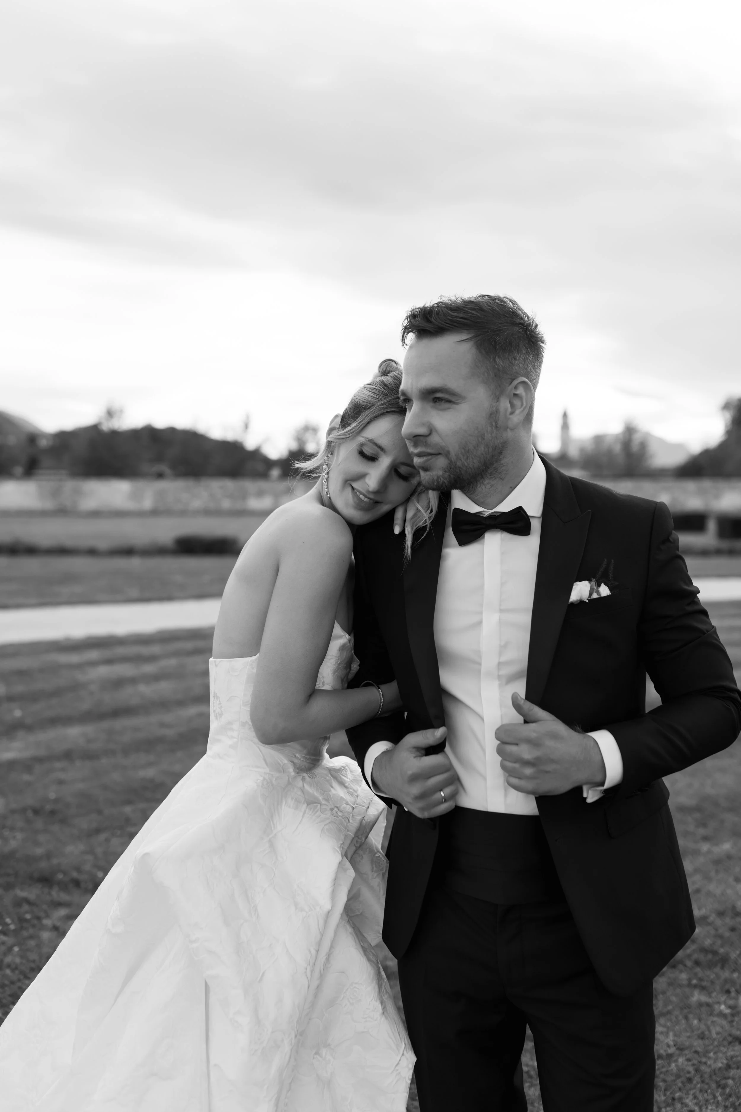 Ein Brautpaar bei einer Hochzeitsaufnahme im Freien, der Frau lehnte sich an den Mann, beide tragen formelle Hochzeitskleidung, der Mann trägt einen schwarzen Anzug und Fliege, die Frau trägt ein weißes Brautkleid.