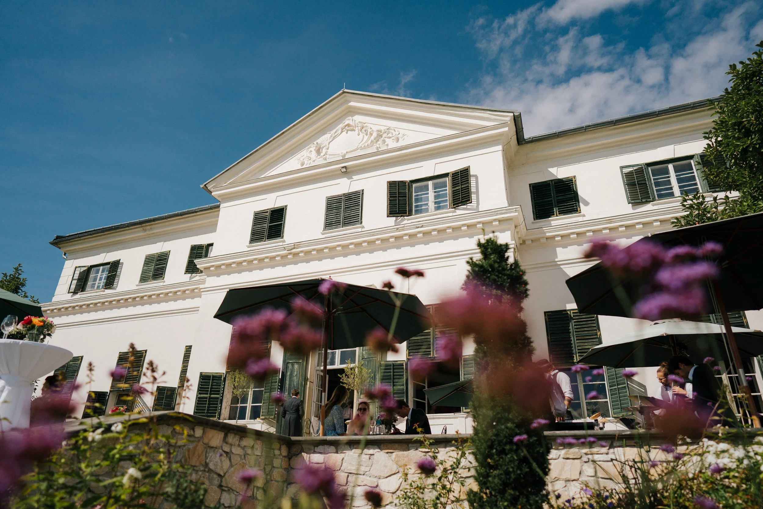 Ein weißes Gebäude mit grünen Fensterläden und einem geschnitzten Giebel unter einem blauen Himmel, im Vordergrund blühende Blumen und Menschen bei einem Gartenfest. Es ist das Schloss Rosegg in Kärnten.