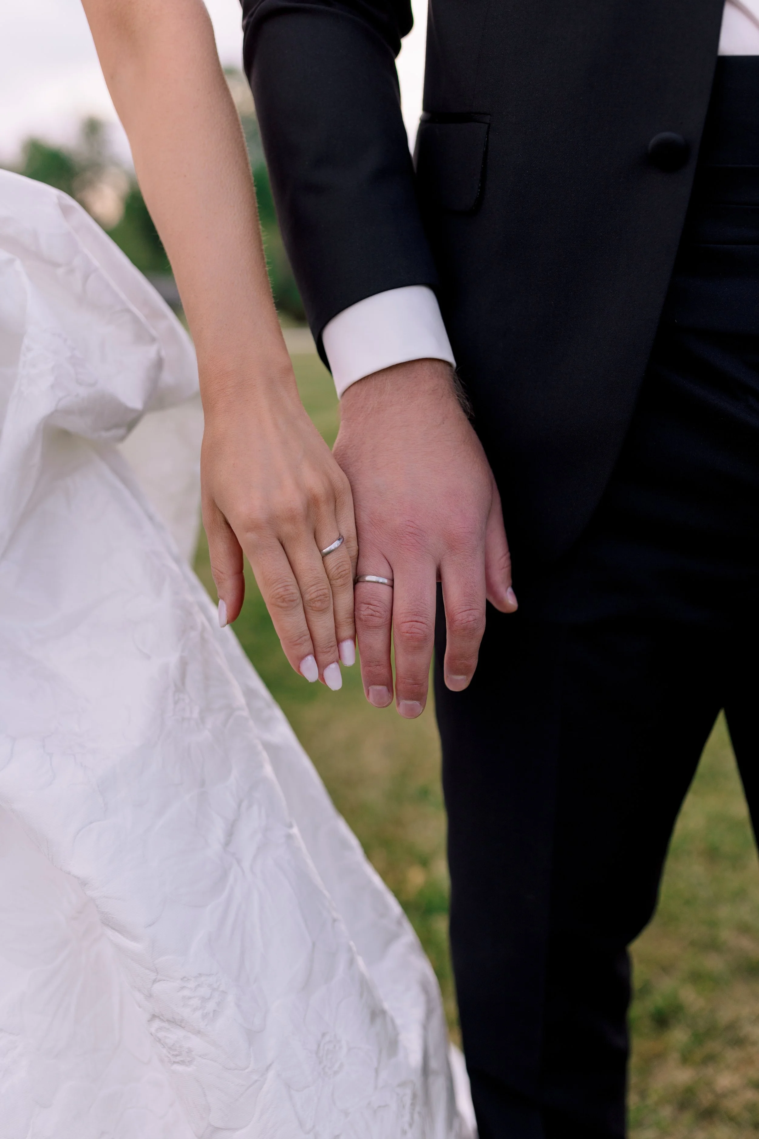 Das Bild zeigt ein Paar, das sich die Hände hält, bei ihrer Hochzeit. Die Frau trägt ein weißes Kleid, und der Mann trägt einen schwarzen Anzug. Beide haben Eheringe am Finger. Der Hintergrund ist unscharf und zeigt eine grüne, natürliche Umgebung.