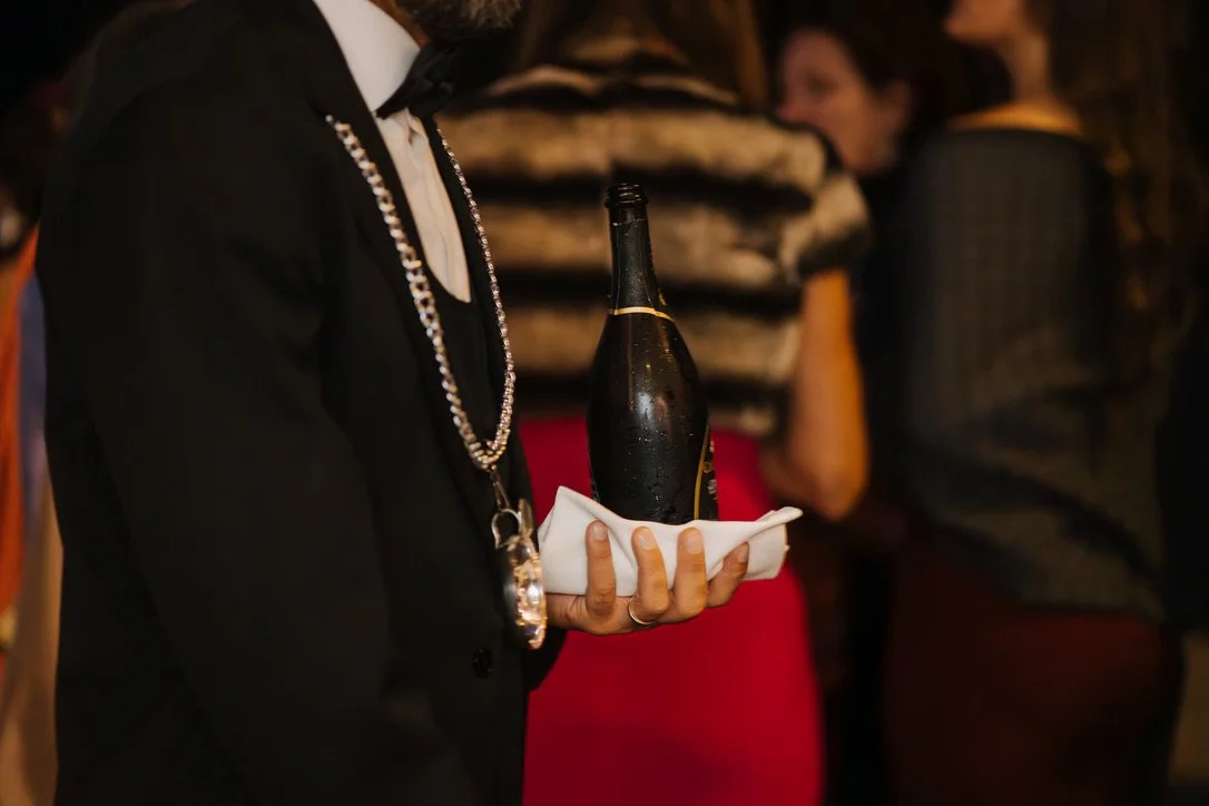 Person in formeller Kleidung hält eine Flasche Champagner auf einem Tablett bei einer Feier.