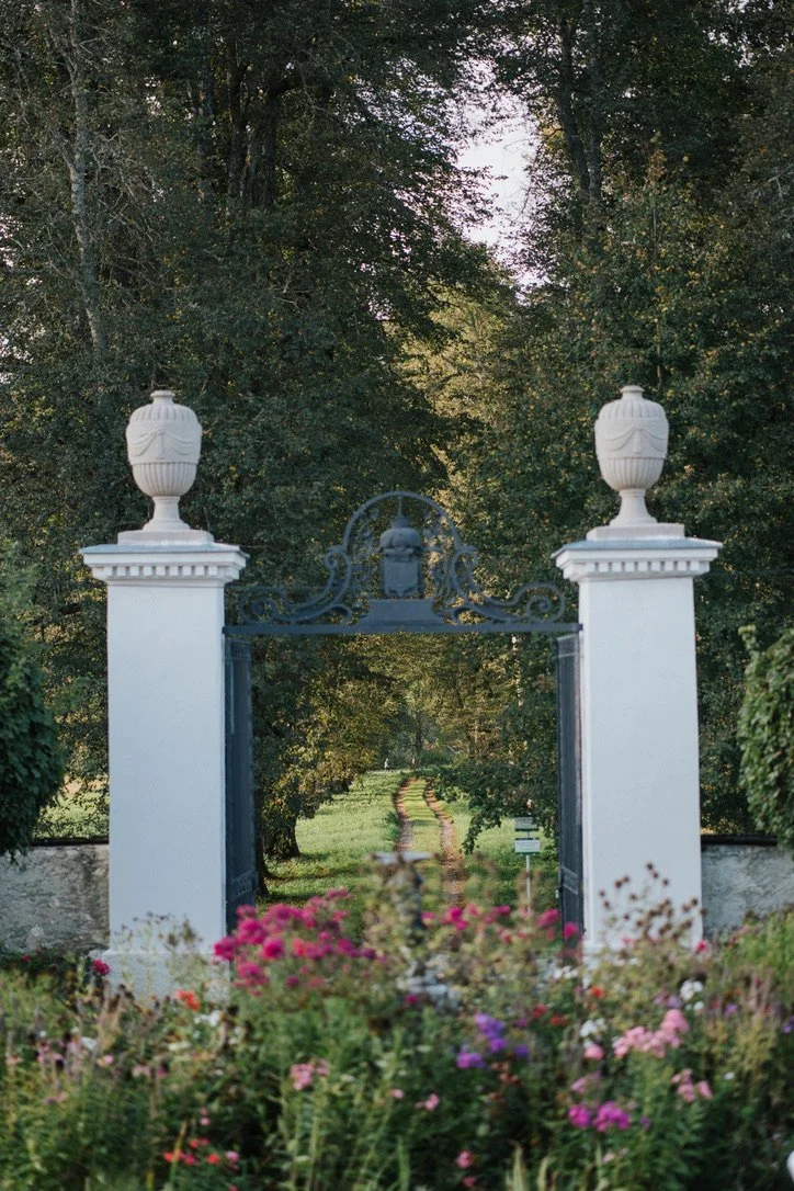 Ein Eingangstor zu einem Garten mit weißen Säulen, blühende Blumen im Vordergrund und einen Weg, der ins Grüne führt, umgeben von Bäumen. Es ist das Tor zum Schloss Rosegg.