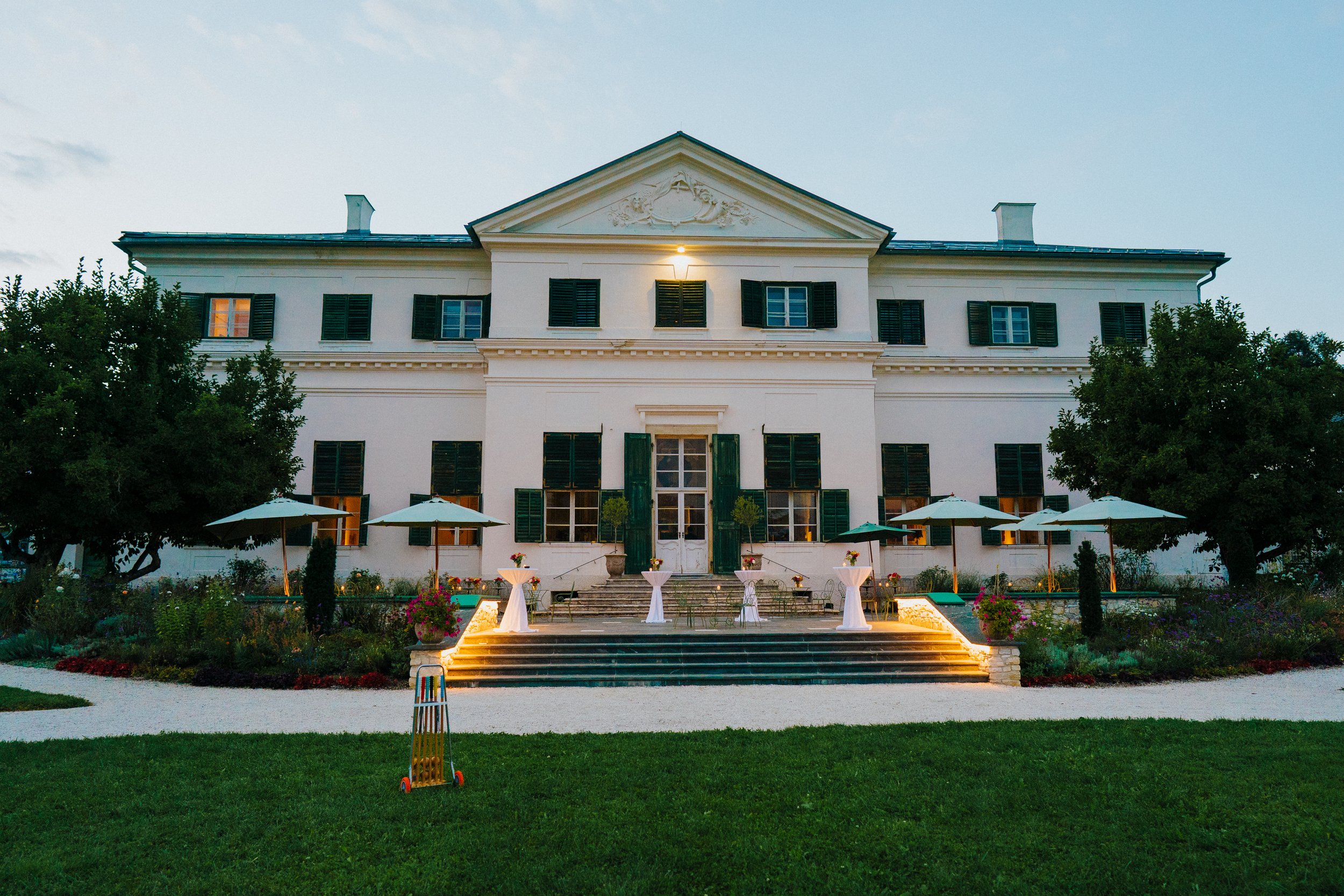 Ein weißes Herrenhaus, Schloss Rosegg mit grünen Fensterläden, umgeben von Bäumen und Gartenmöbeln bei Dämmerung, beleuchtet mit Lampen im Vorgarten.