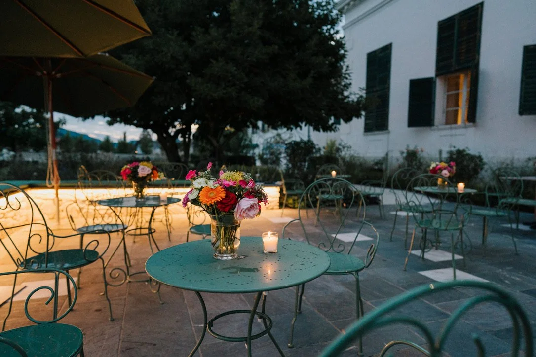 Terrasse von Schloss Rosegg, ein Außenbereich mit mehreren Tischen und Stühlen, dekoriert mit Blumensträußen und Kerzen, bei Dämmerung.