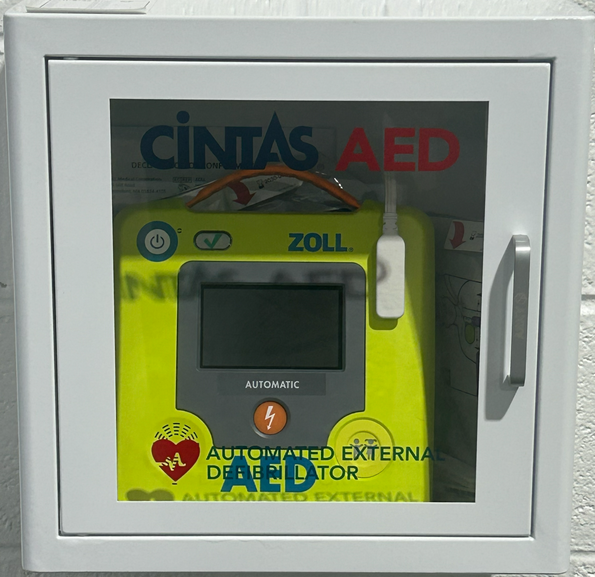 Automated external defibrillator (AED) inside a white cabinet with a glass door, labeled 'CINTAS AED' on top. The AED is yellow with a small screen, power button, and a white cable connected to its side.