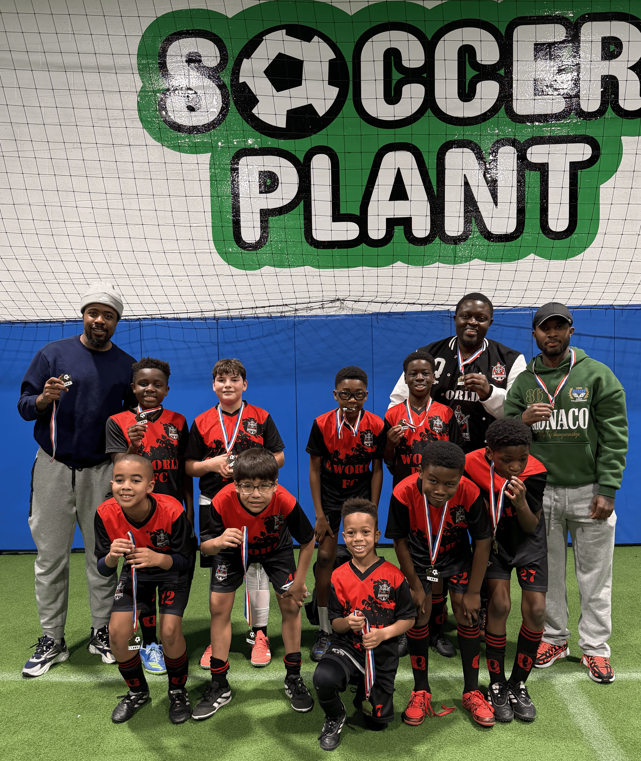 Group of young soccer players and coaches posing with medals in an indoor soccer facility with a large 'Soccer Planet' sign overhead.