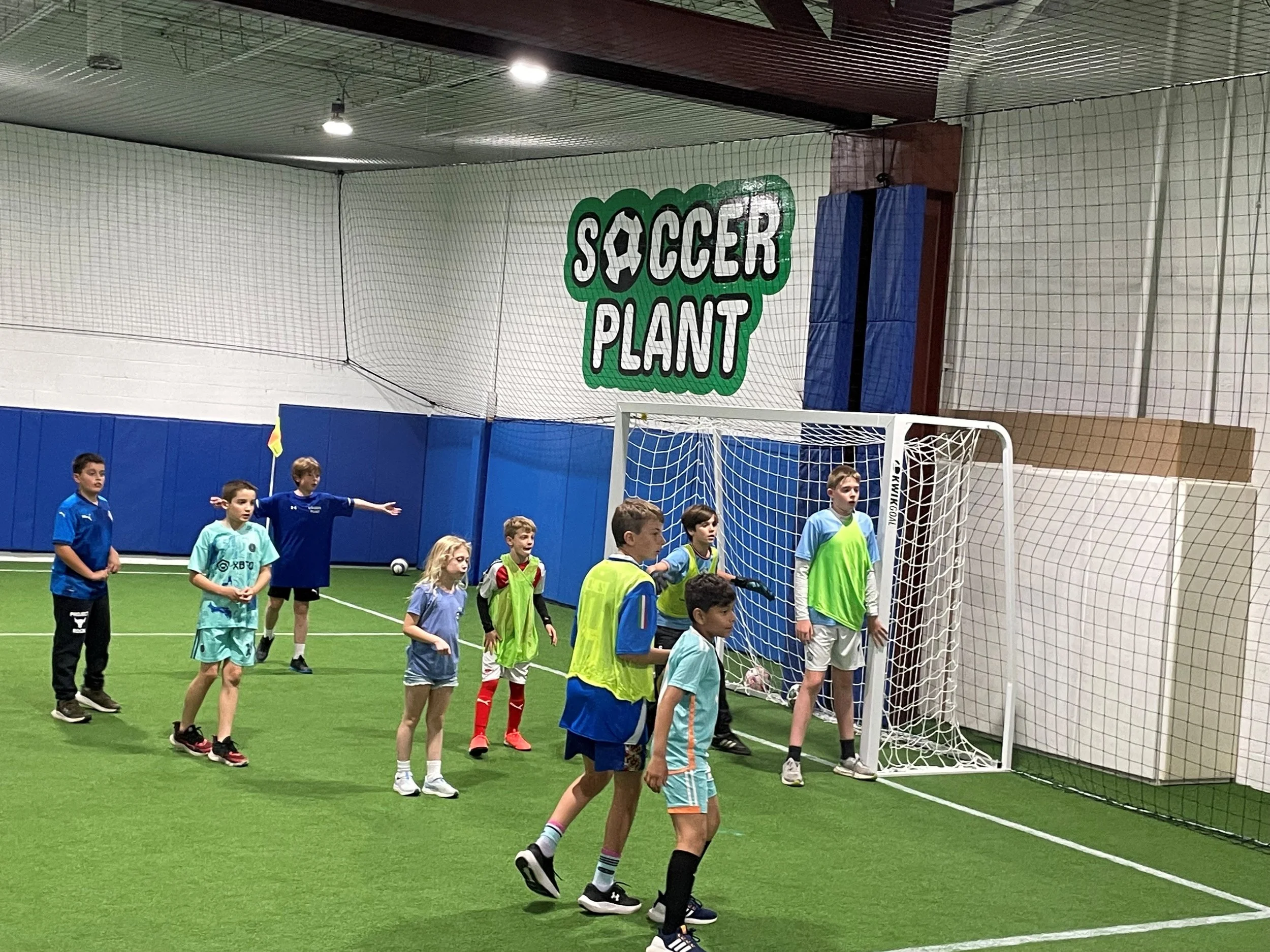 Children playing indoor soccer on a green turf field, with a sign saying 'Soccer Plant' on the wall.