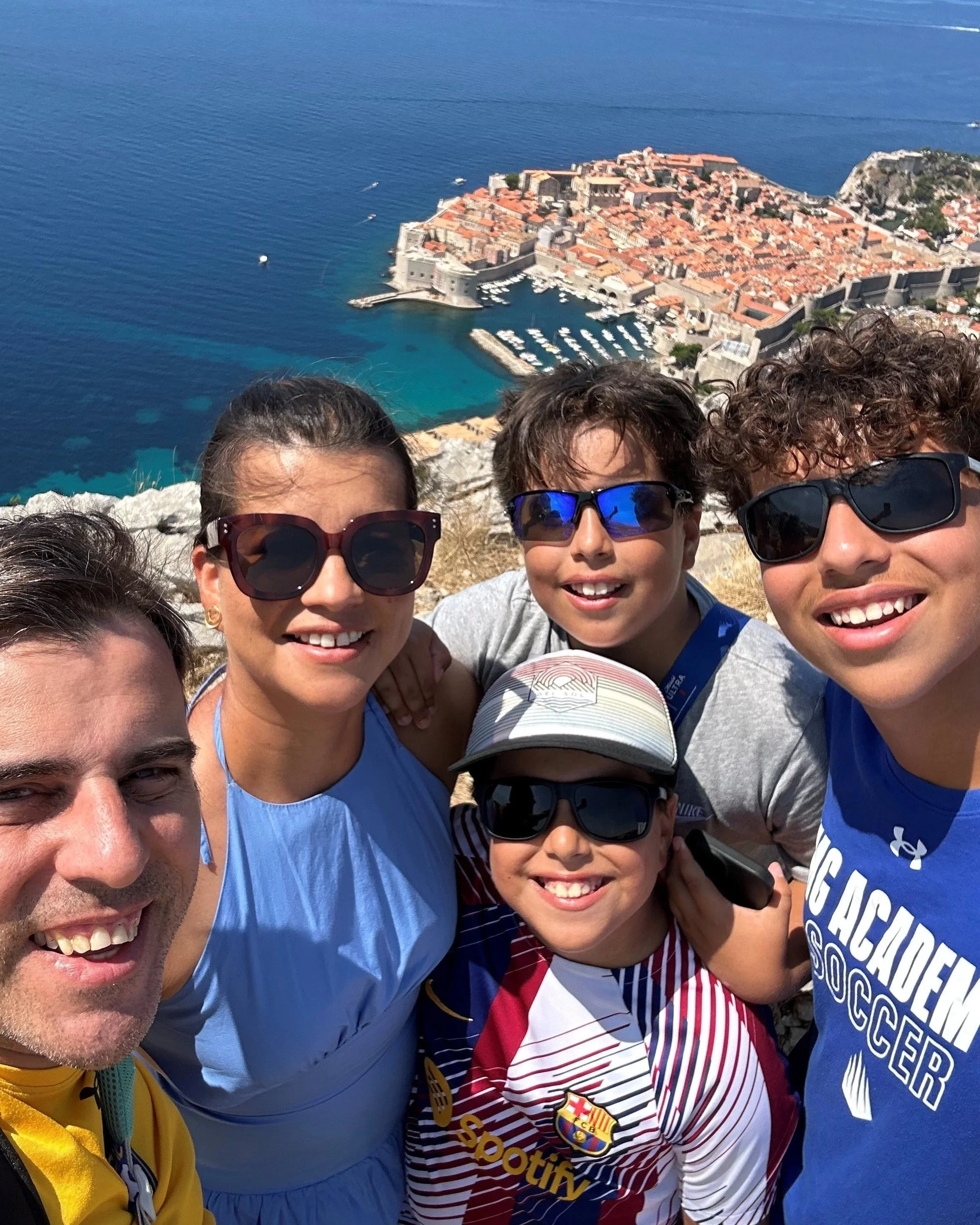 A group of five people taking a selfie on a hillside with a coastal town and harbor in the background.