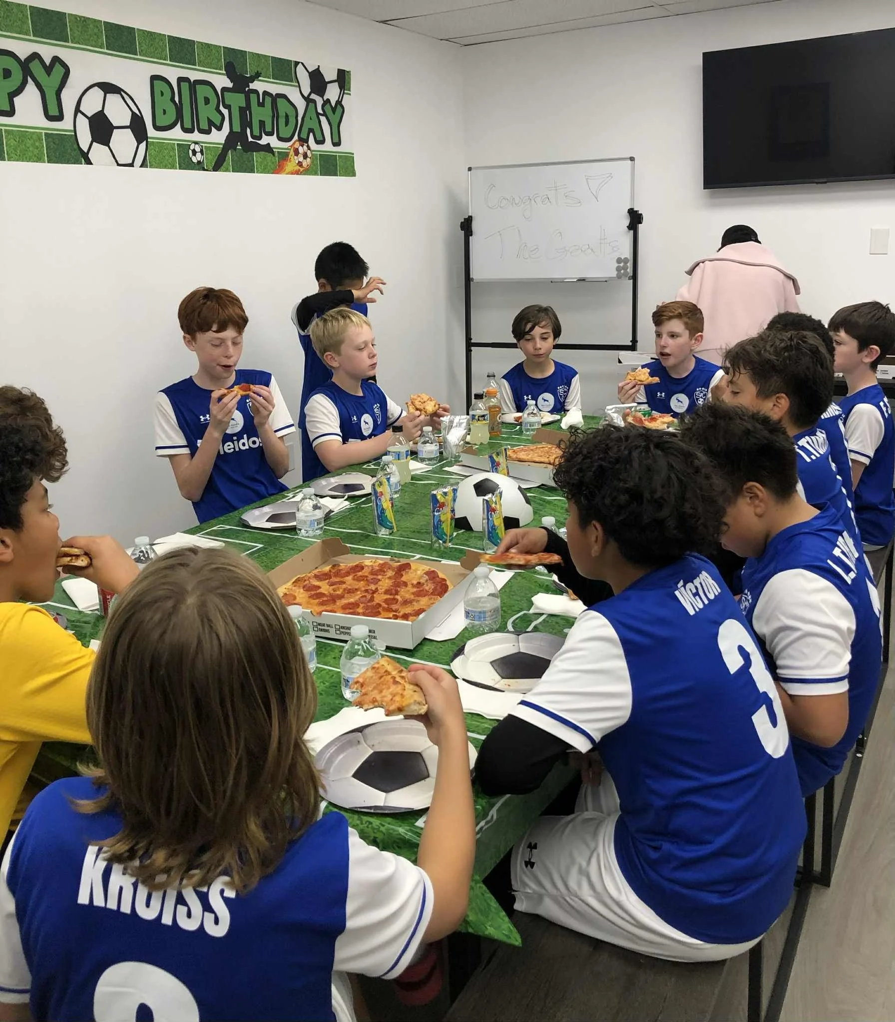 Kids in blue soccer jerseys at a birthday party eating pizza around a decorated table.