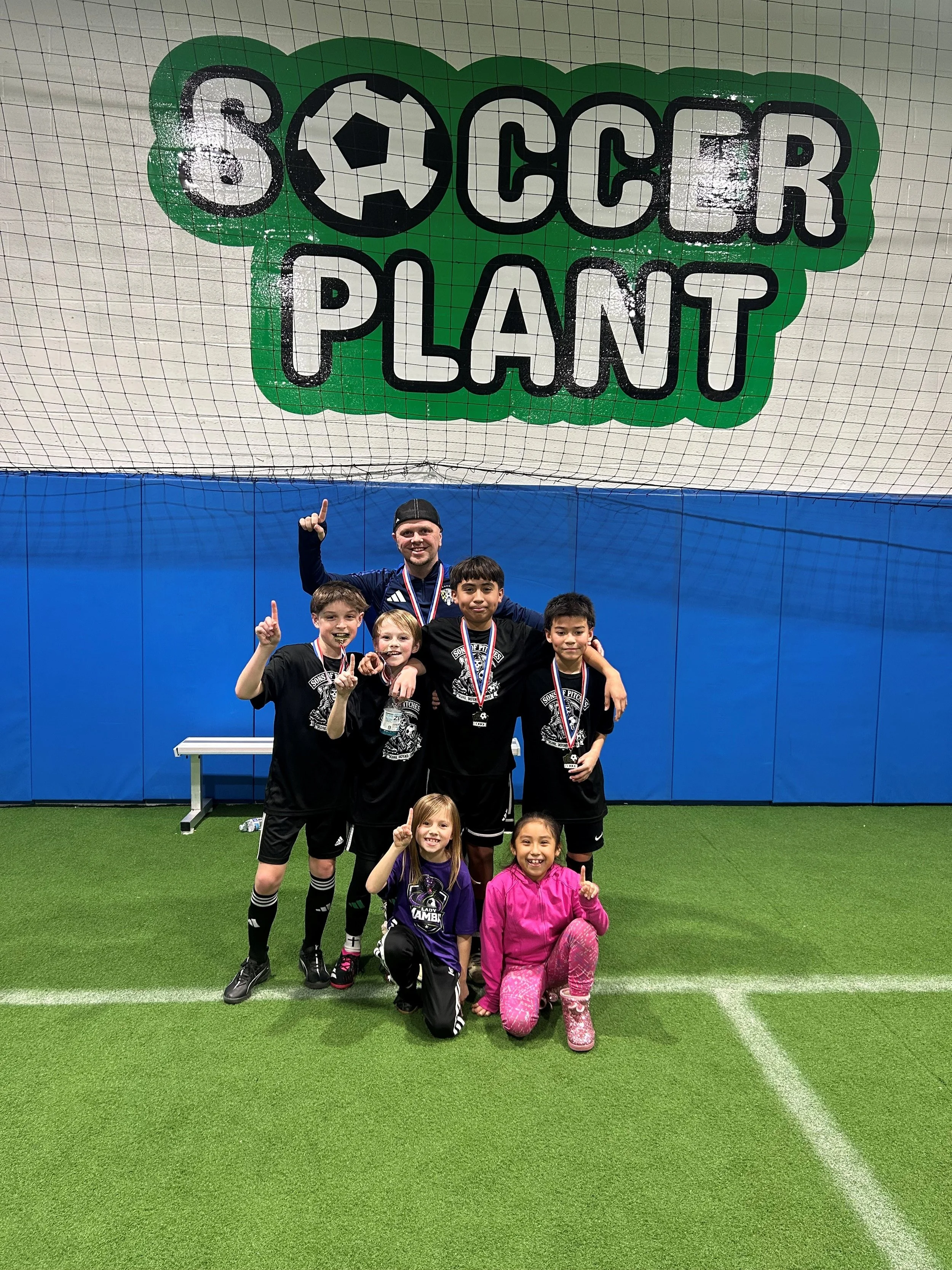 A group of children and a coach are celebrating after a soccer game, standing on an indoor soccer field with a blue wall, where a large sign reading