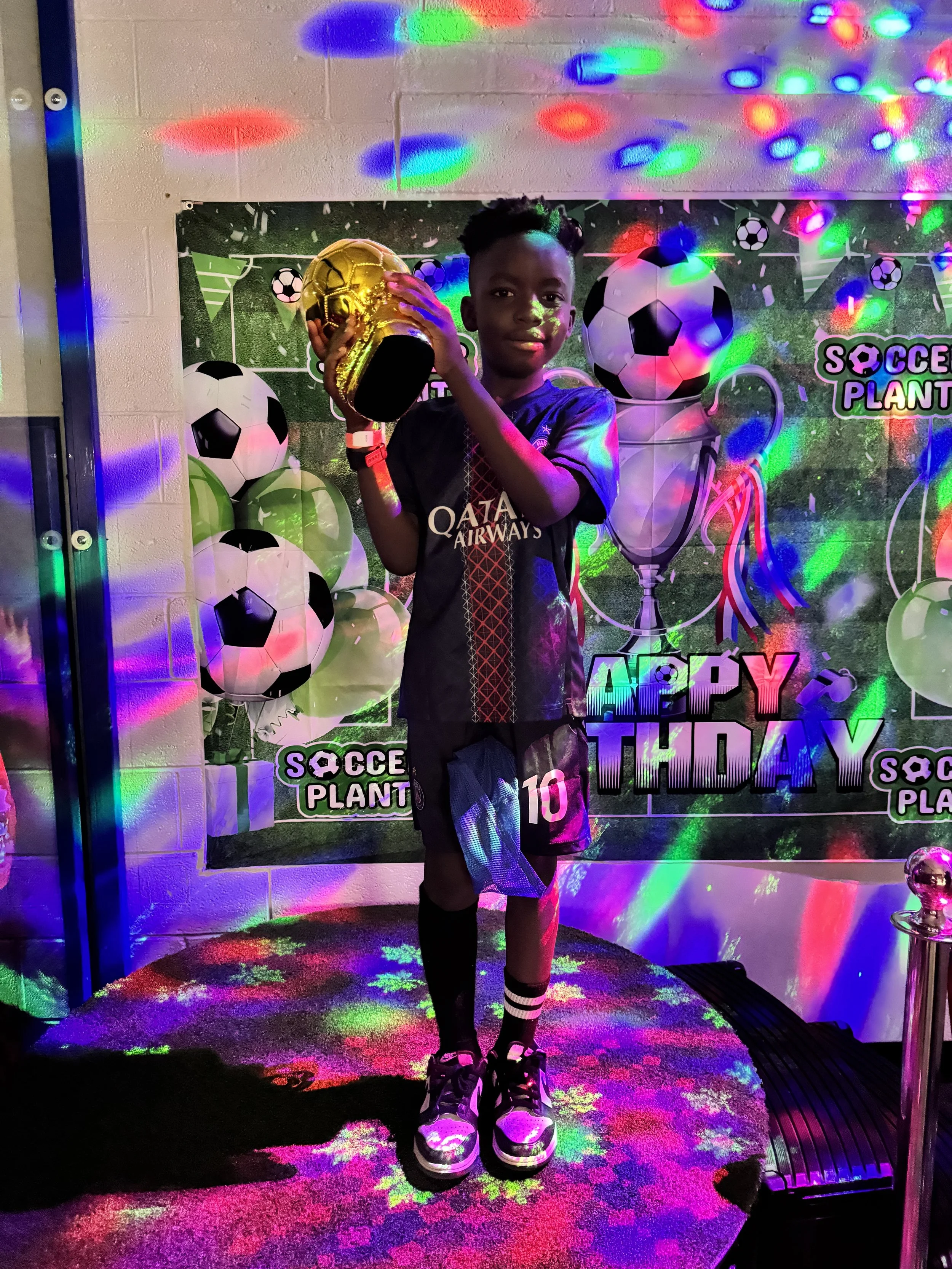 A young boy in a soccer jersey posing with a golden soccer ball in front of a birthday backdrop with soccer-themed decorations, colorful lights, and the words 'Happy Birthday'.