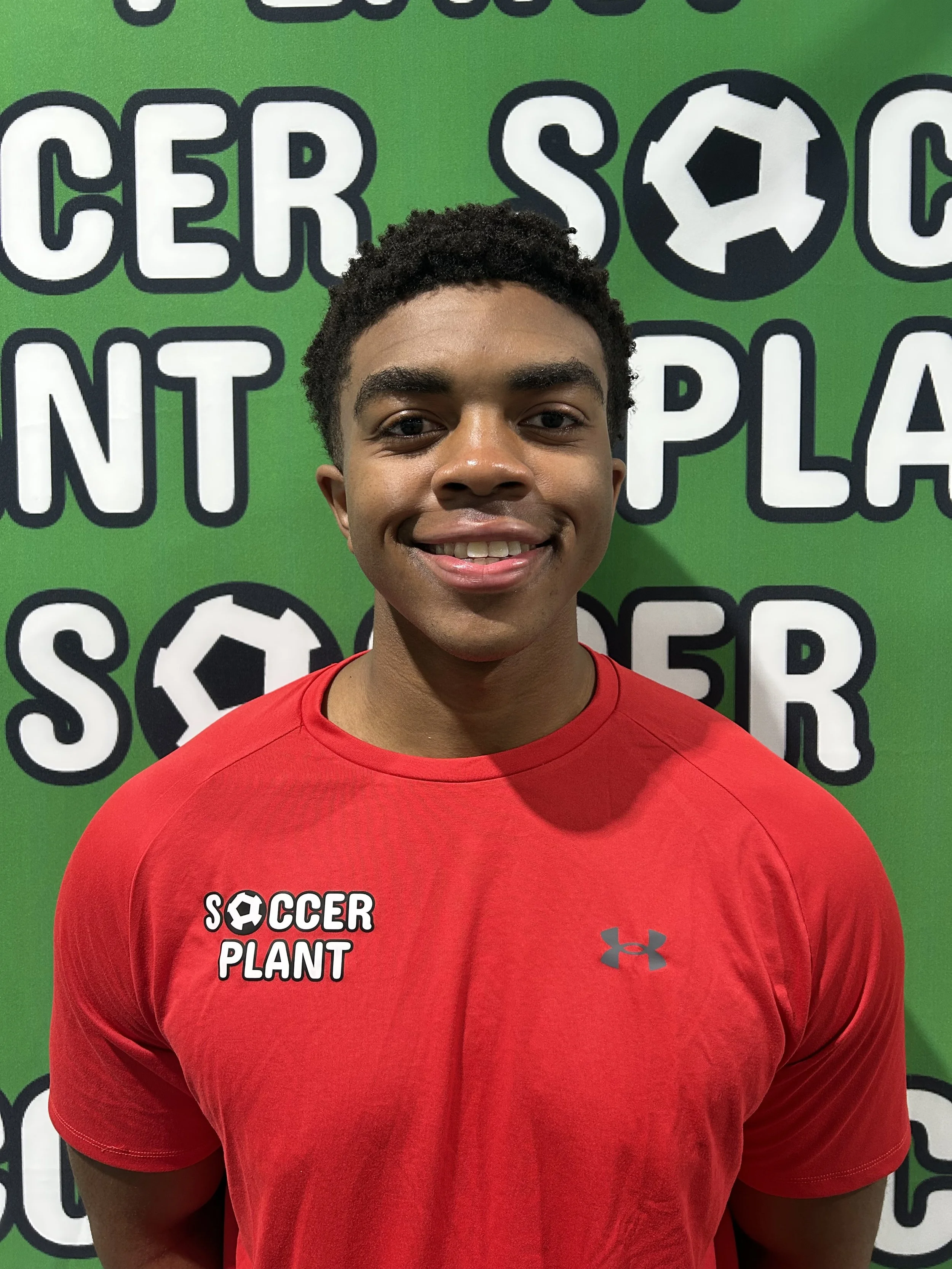 Young man smiling, wearing a red athletic shirt with 'Soccer Plant' logo, standing in front of a green background with 'Soccer Plant' logo and text.