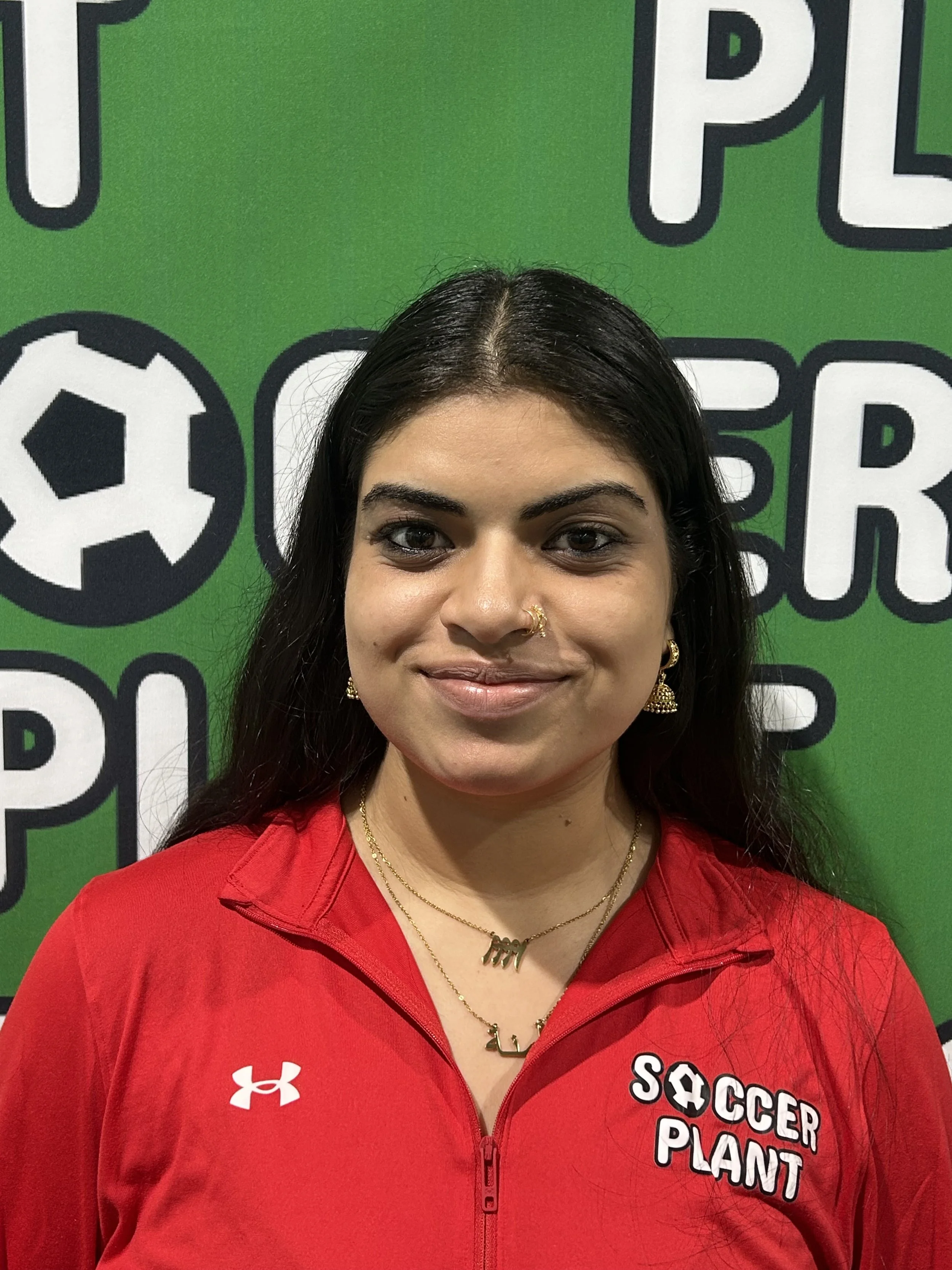Close-up of a woman with dark hair and earrings, wearing a red jacket with 'Soccer Plant' logo, standing in front of a green background with sports-themed signage.