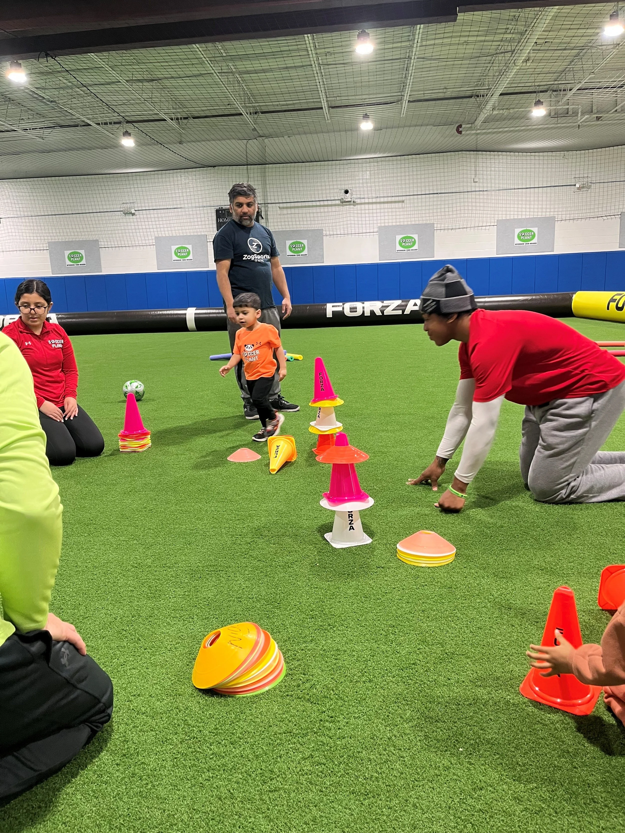 Children and adults participating in an indoor soccer drill with cones and flags on artificial turf.
