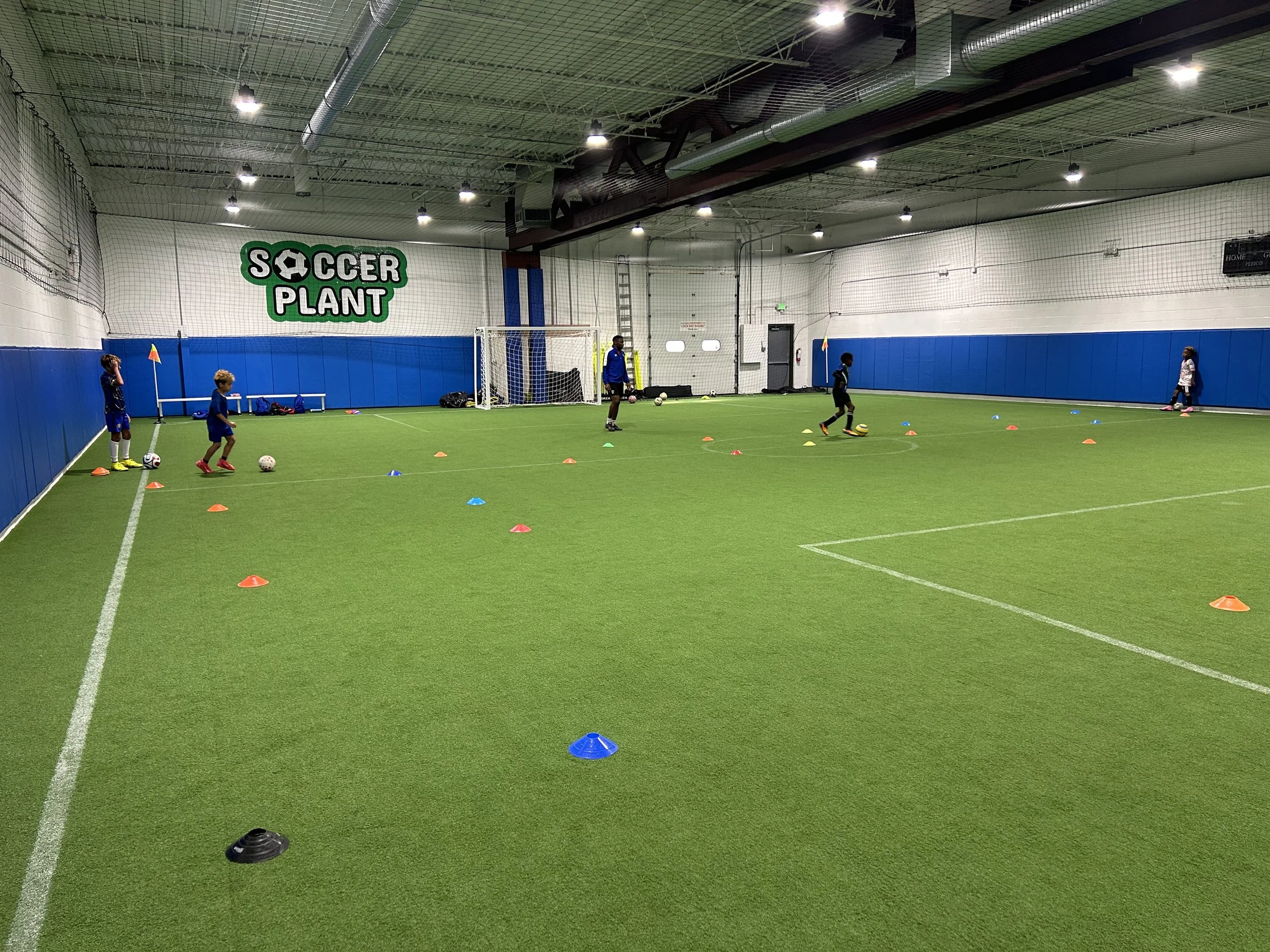 Indoor soccer training session with young players practicing dribbling and shooting, marked by colorful cones on a synthetic turf field, with a goal, nets, and a sign that reads 'Soccer Plant' in the background.