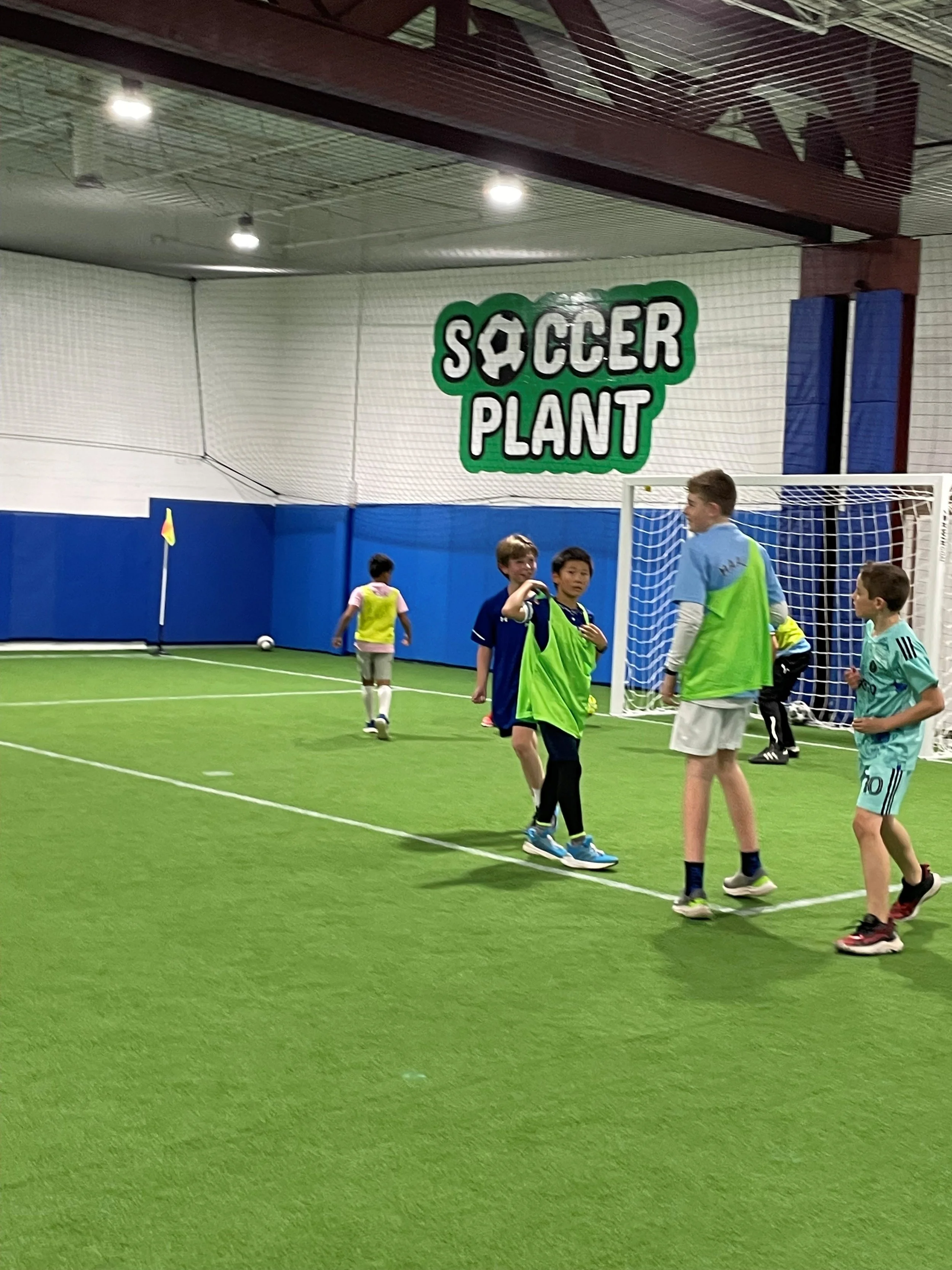 Children playing soccer inside an indoor soccer facility called Soccer Plant, with a goal and a corner flag visible.