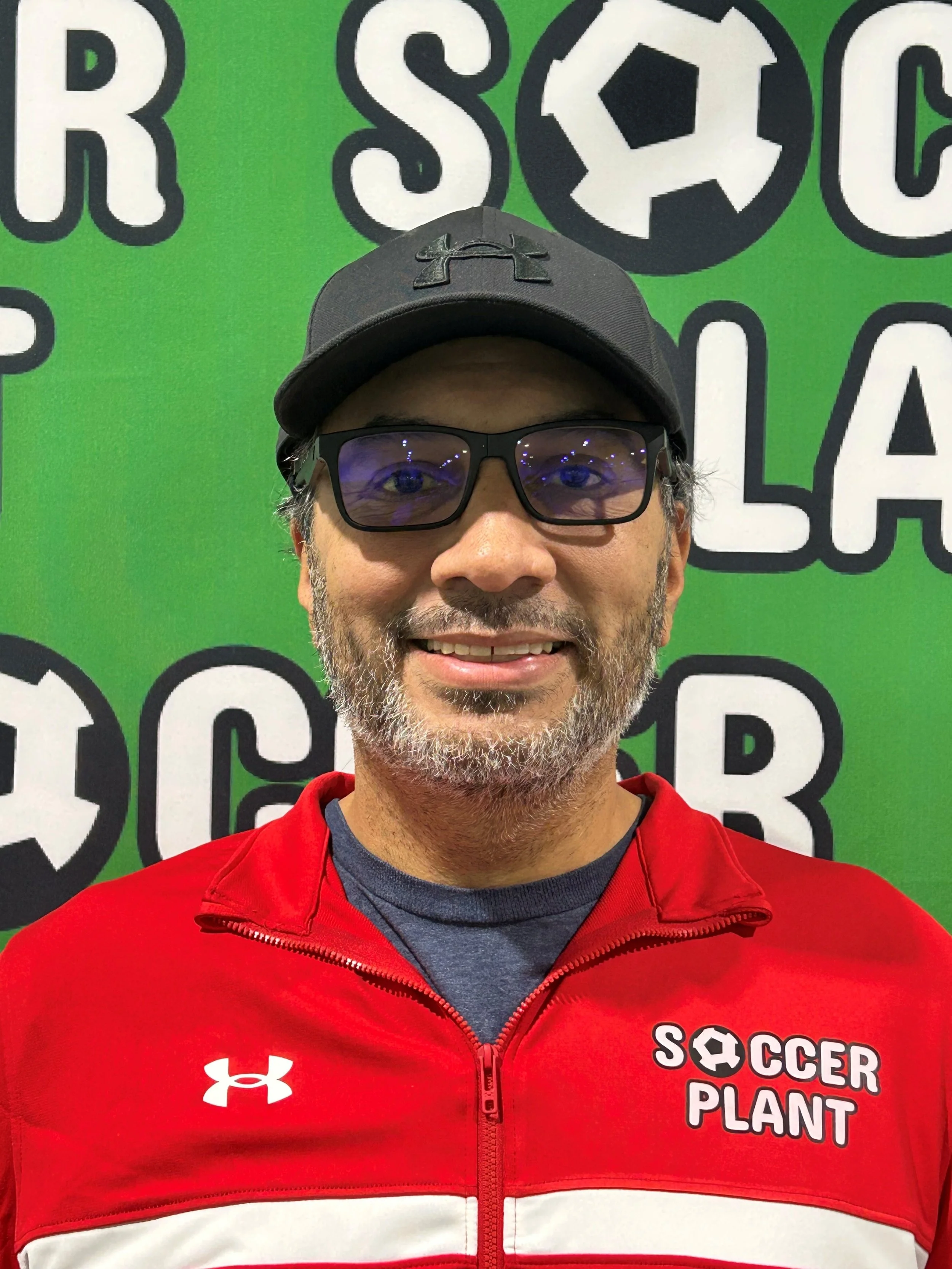 A man wearing a black cap, black glasses, a red Under Armour jacket with the words 'Soccer Plant' on it, standing in front of a green background with soccer-themed logos.