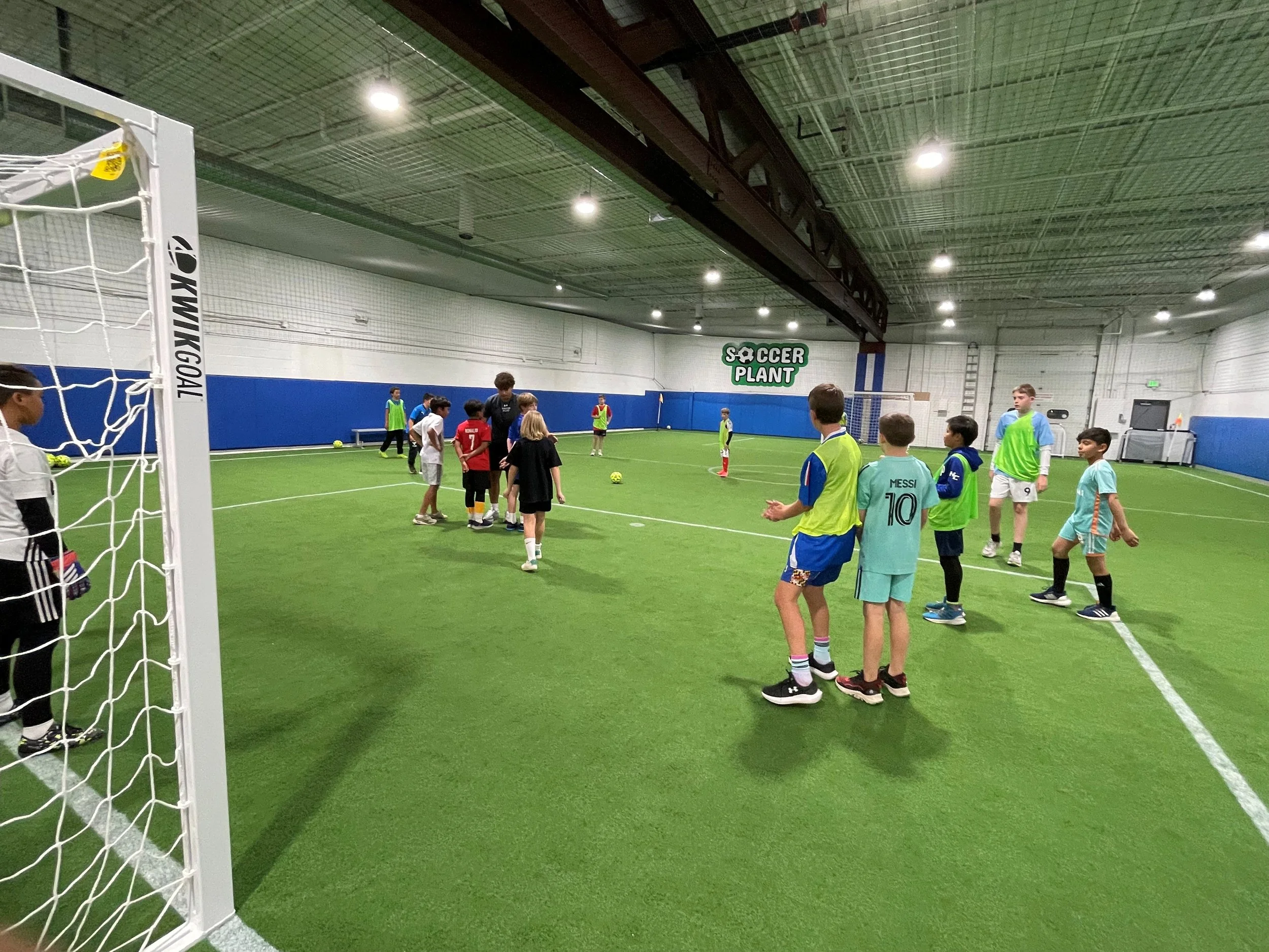 Students practicing soccer on an indoor field during a training session, with kids and coaches. The field has a goalpost, artificial turf, and a sign in the background that says 'Soccer Plant'.
