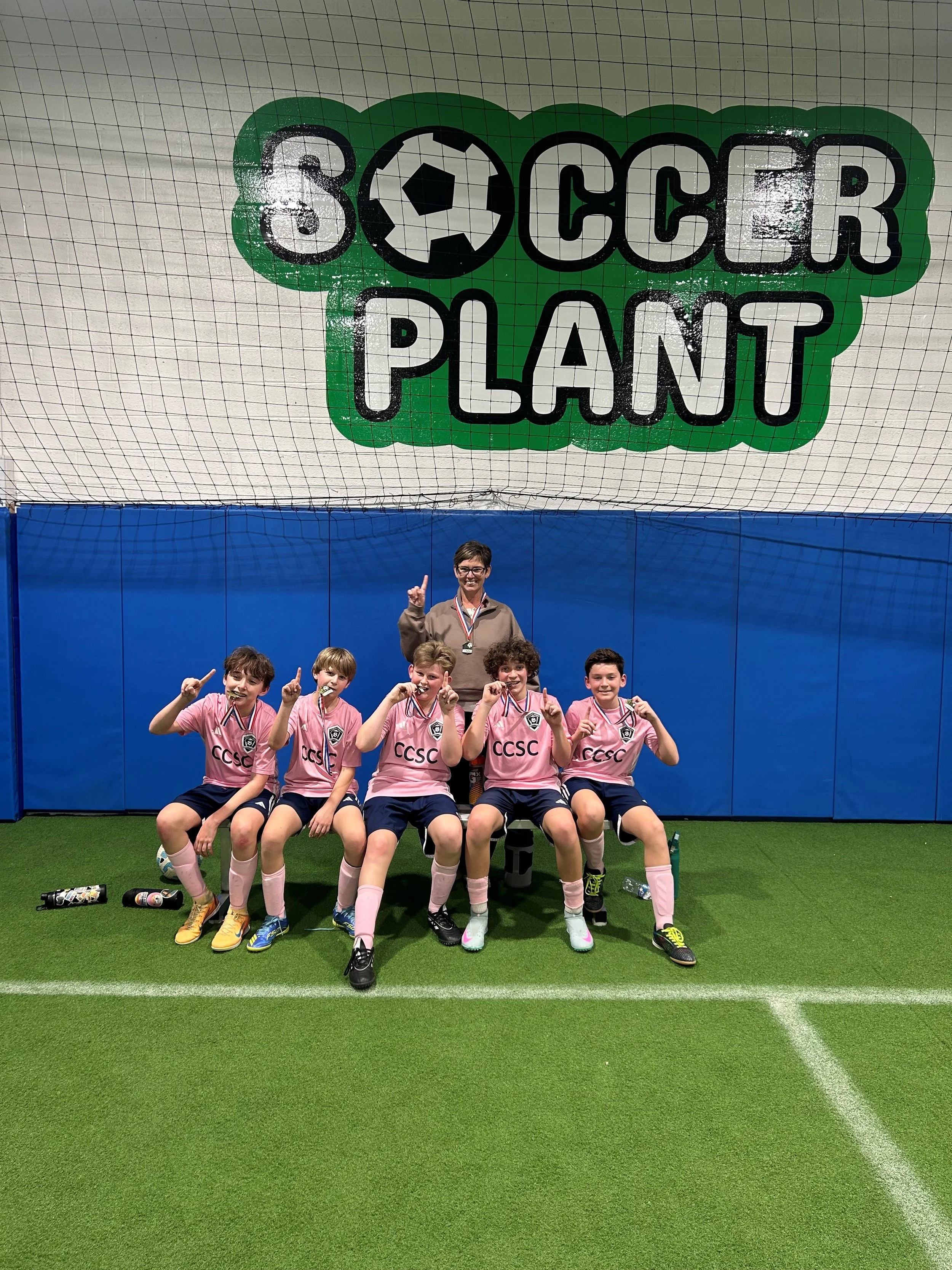 Group of young female soccer players in pink uniforms sitting on the field with their coach, all wearing medals, inside an indoor soccer facility with a large sign saying 'Soccer Planet' above them.
