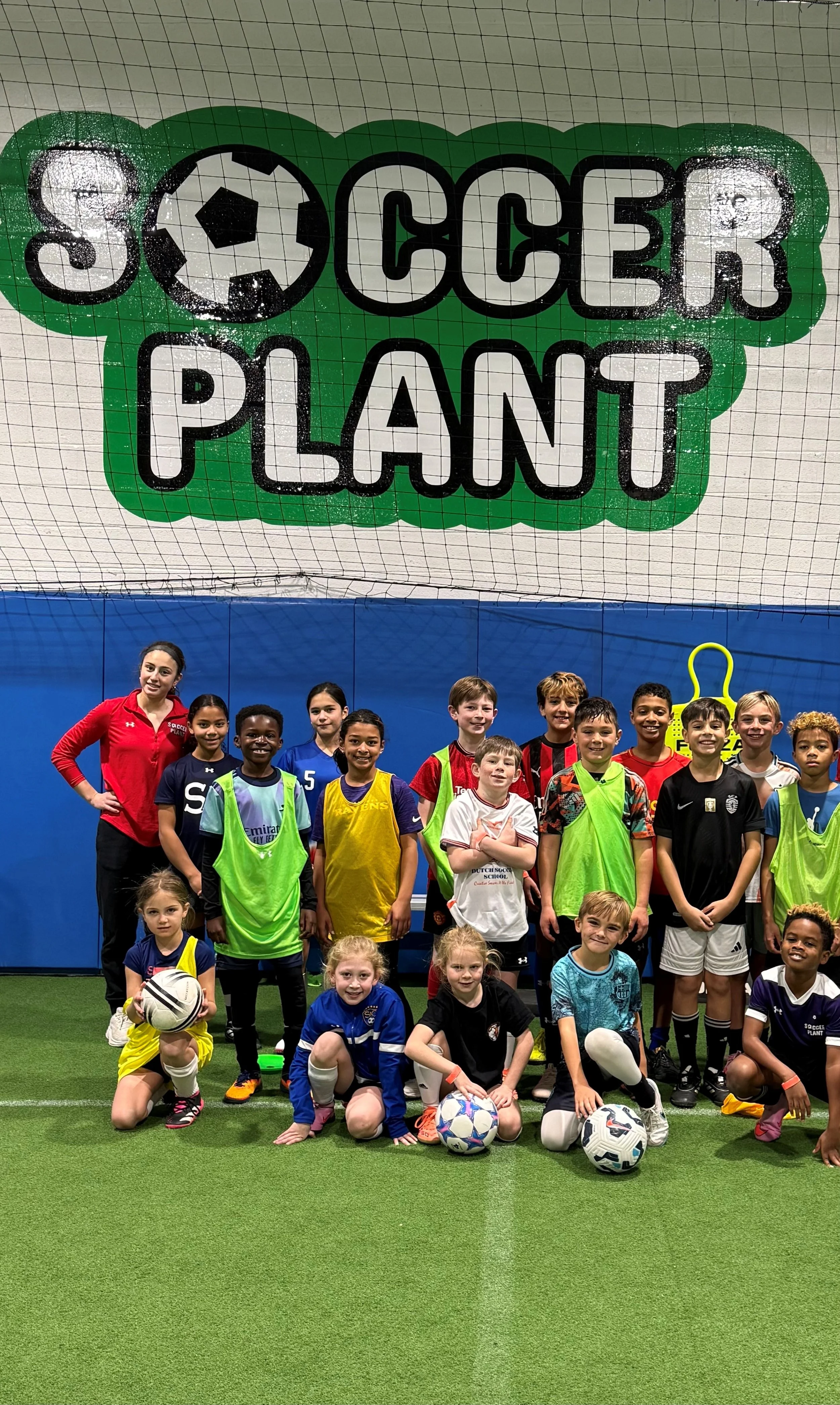 Group of children and an adult coach in soccer uniforms posing for a team photo inside an indoor soccer facility with artificial turf and a large 'Soccer Planet' sign featuring a soccer ball graphic in the background.