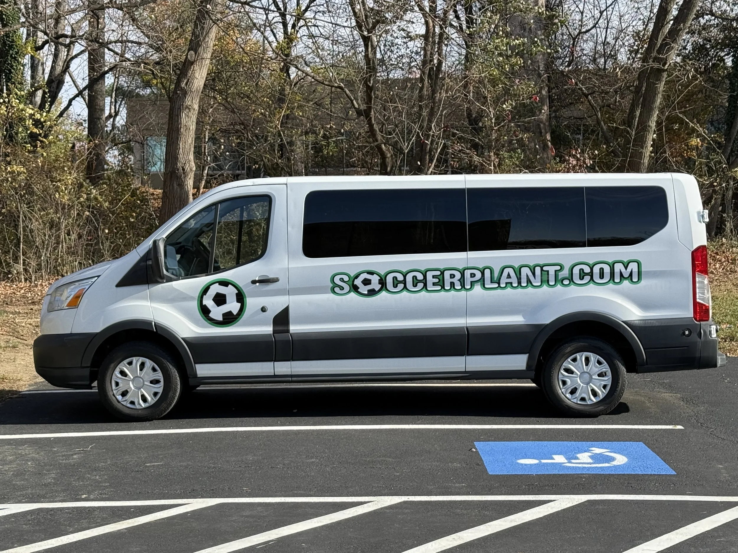 White van with green and black lettering and logo reading 'SOCCERPLANT.COM' parked in a space with a blue handicapped parking sign.