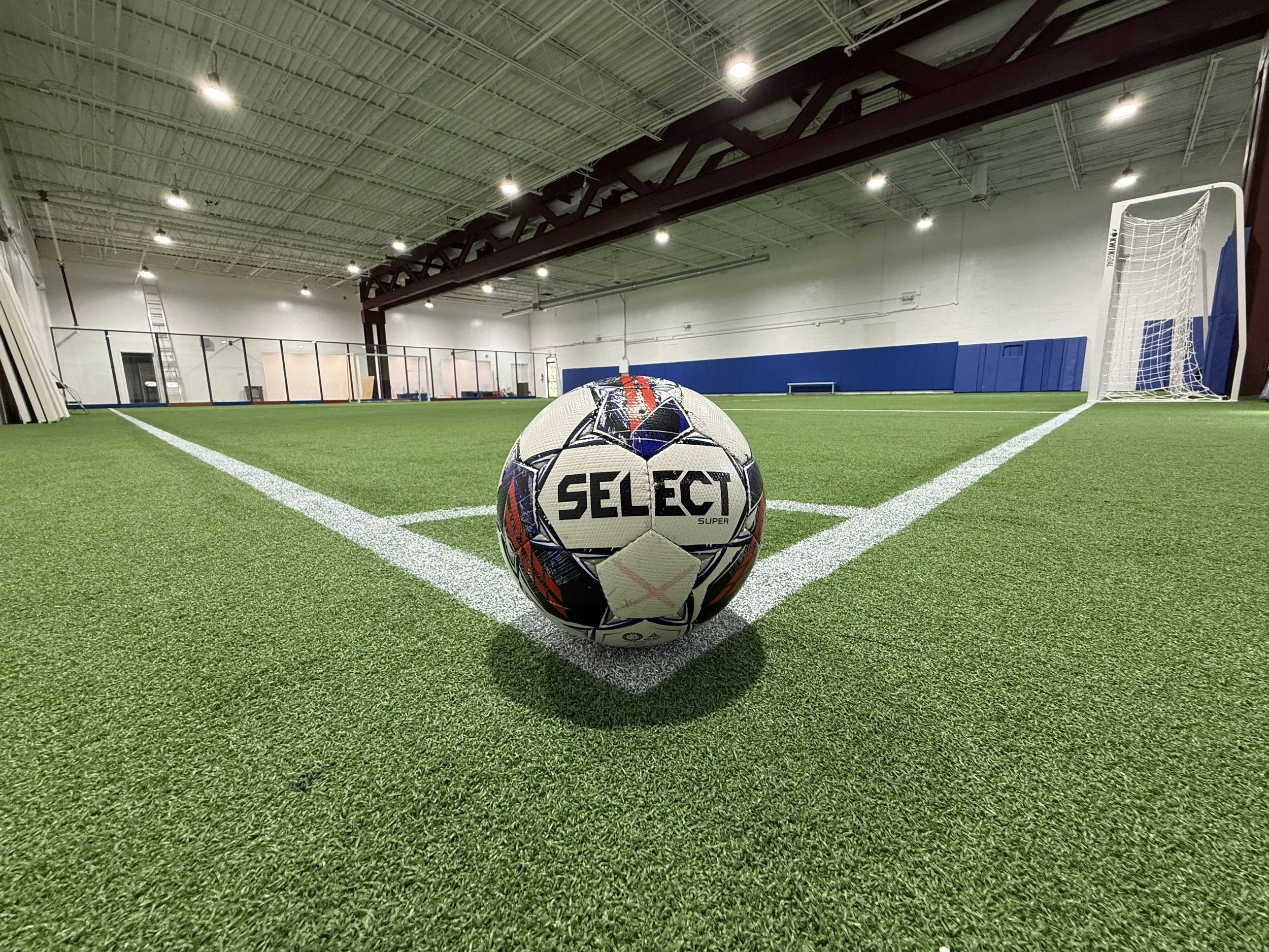 Indoor soccer field with a soccer ball positioned at the corner of the penalty area, goal post in the background, and bright ceiling lights.