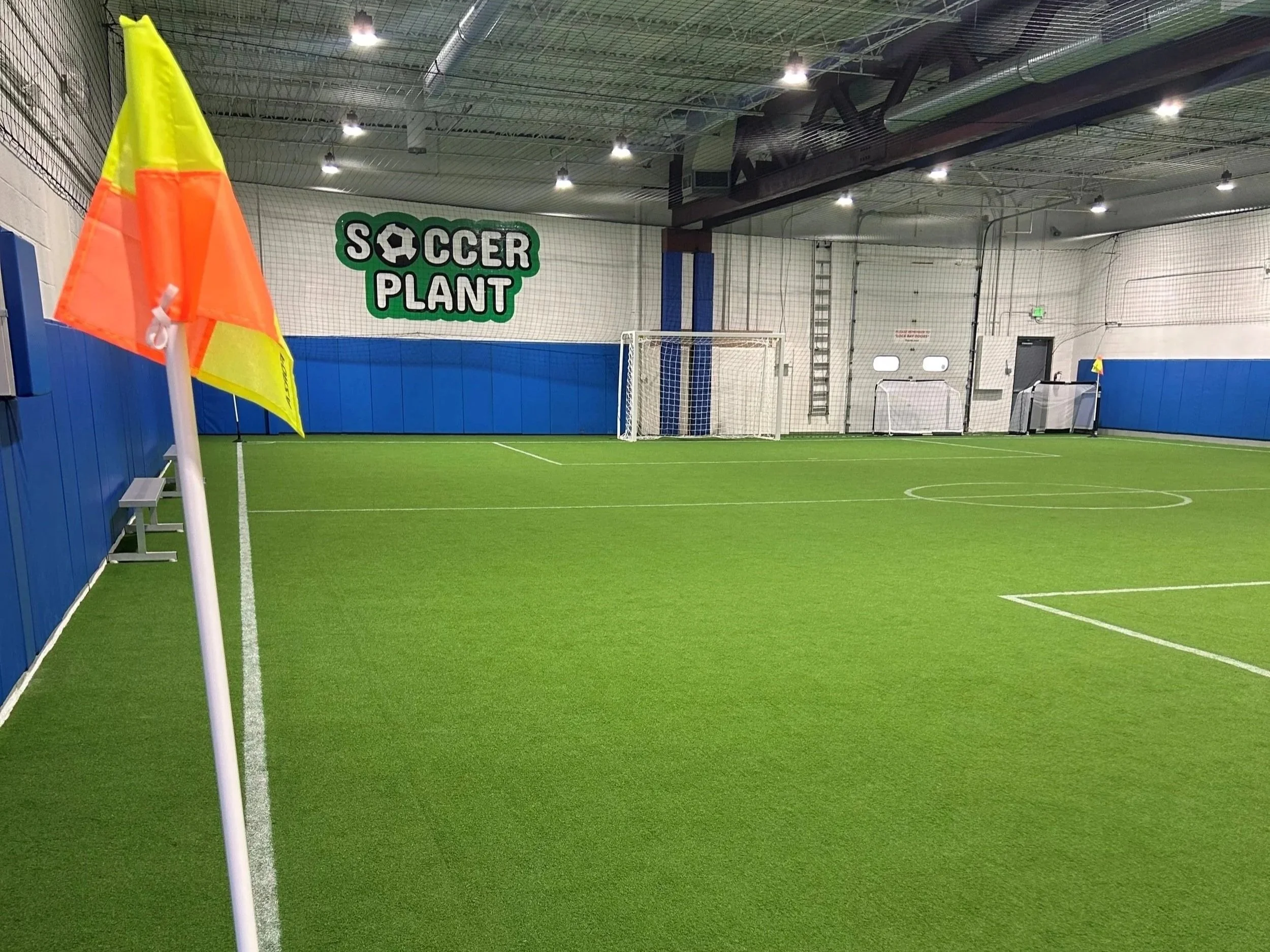 Indoor soccer field with artificial turf, corner flag, goal, and a large sign that says 'Soccer Plant' on the wall.