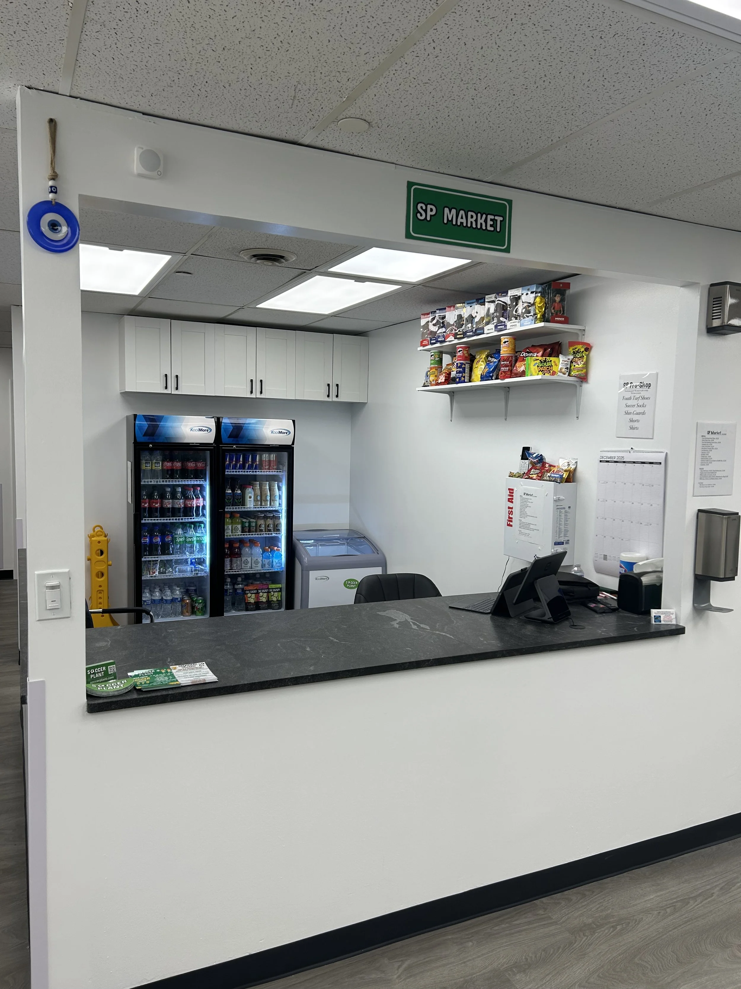 Front counter of a small store or office with a sign reading 'SP MARKET'. Behind the counter are white cabinets, two refrigerator units with beverages, a clothing rack, and a freezer. On the counter, there is a tablet, papers, and a small display.