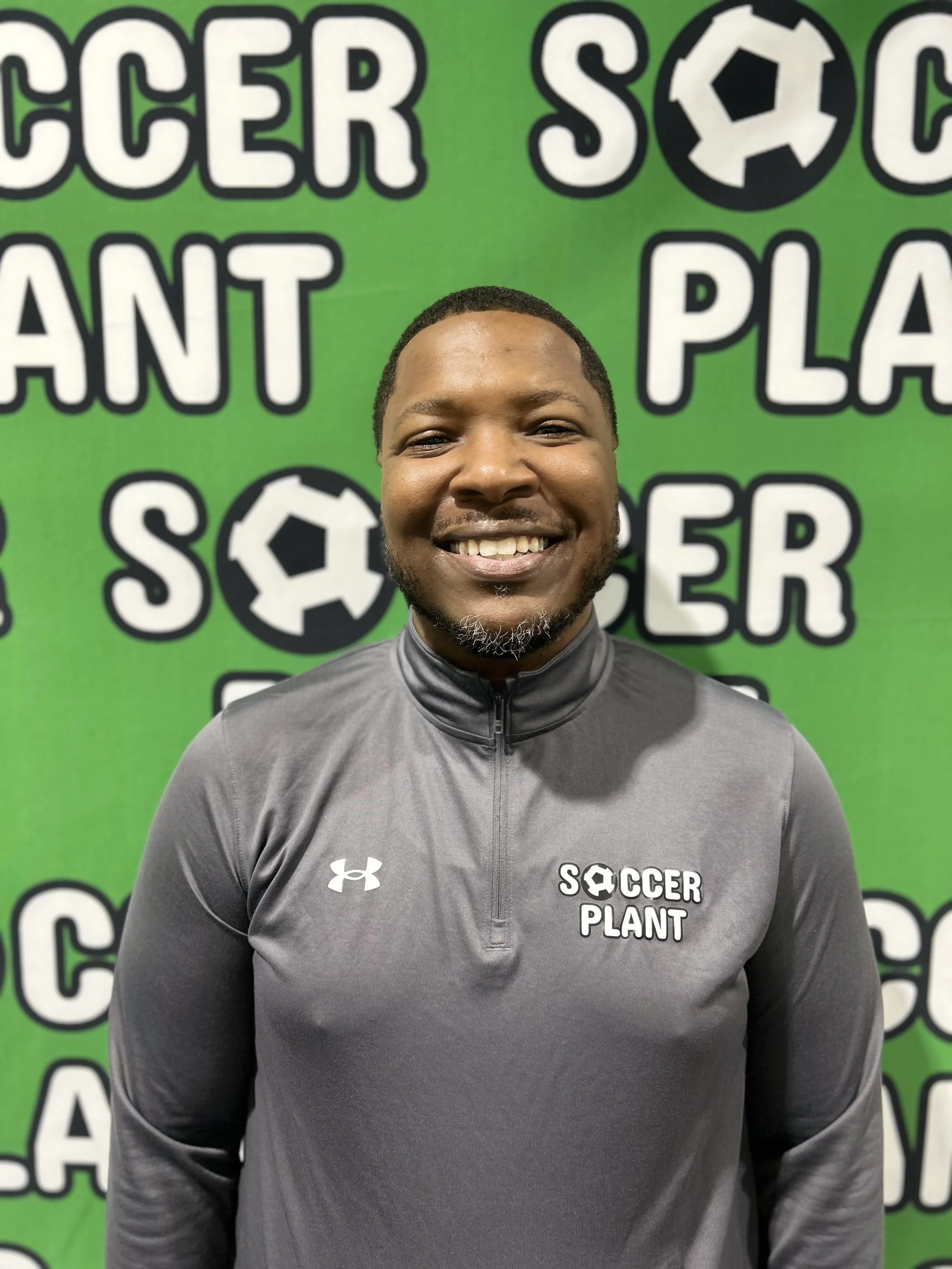 A smiling man standing in front of a bright green background with the words 'Soccer Plant' and soccer ball graphics. He is wearing a gray Under Armour athletic jacket with the logo and 'Soccer Plant' embroidered on it.