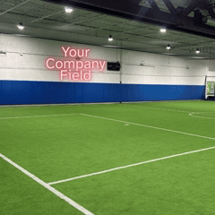 Indoor sports field with artificial grass and white lines, with a sign reading 'Your Company Field' on the wall.