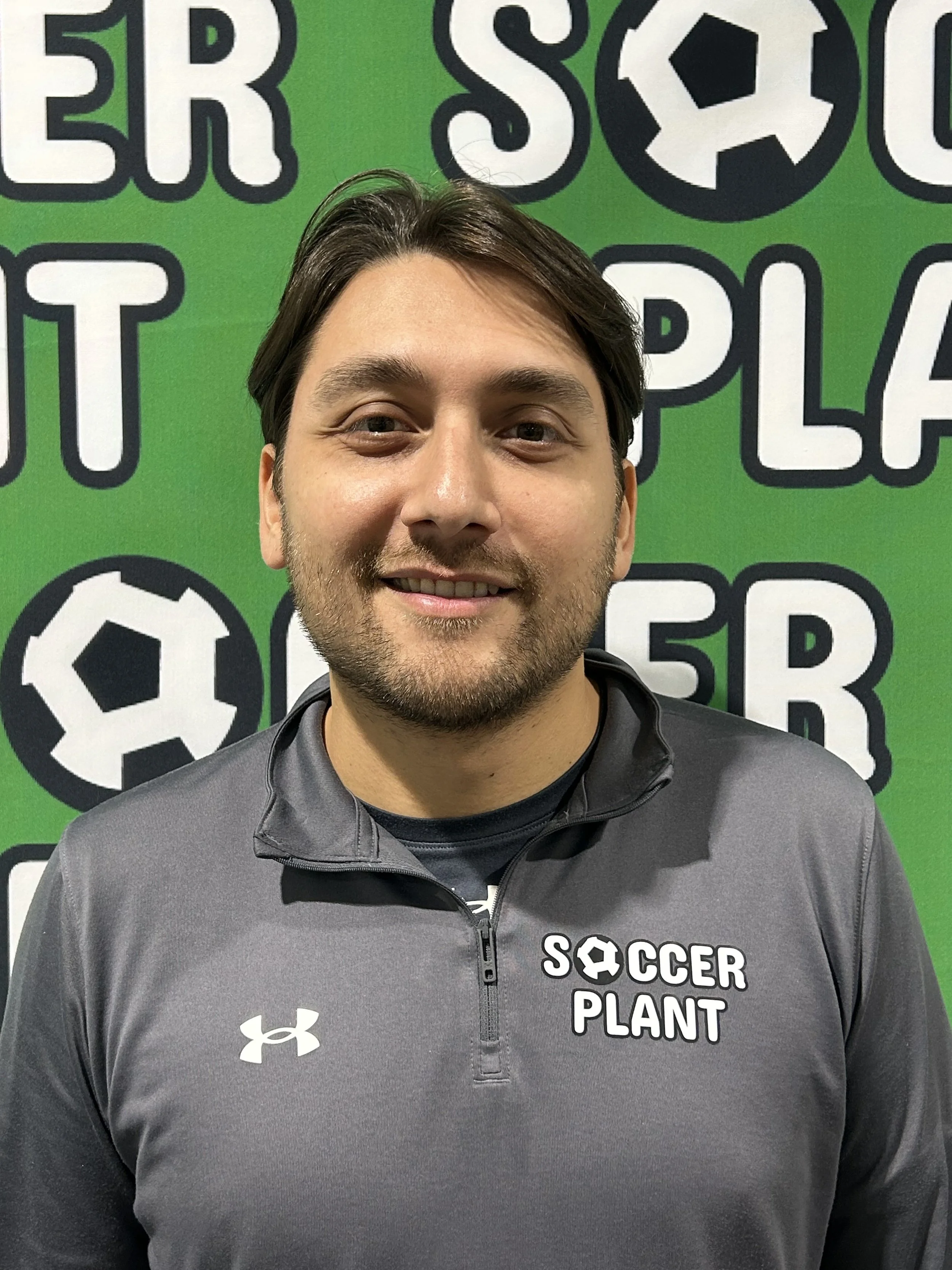 A young man with dark hair and a beard, smiling, wearing a gray Under Armour jacket with the words 'Soccer Plant' and a soccer ball logo on it, standing in front of a green background with soccer-themed text and logos.