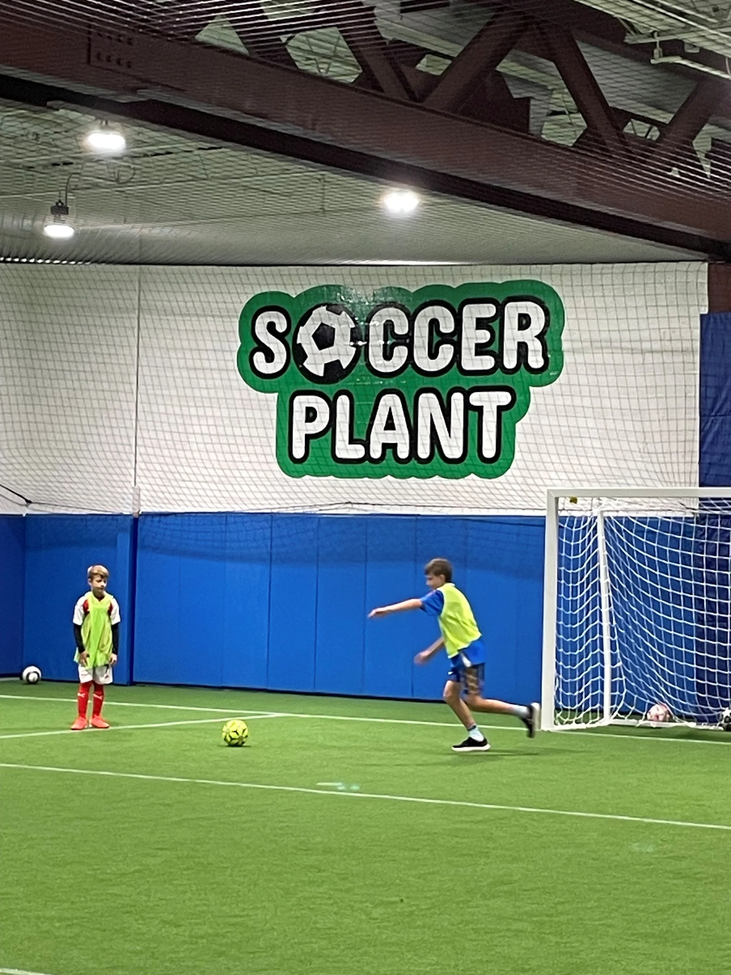 Children playing soccer indoors at soccer training facility with large sign that reads 'Soccer Plant' with a soccer ball logo.
