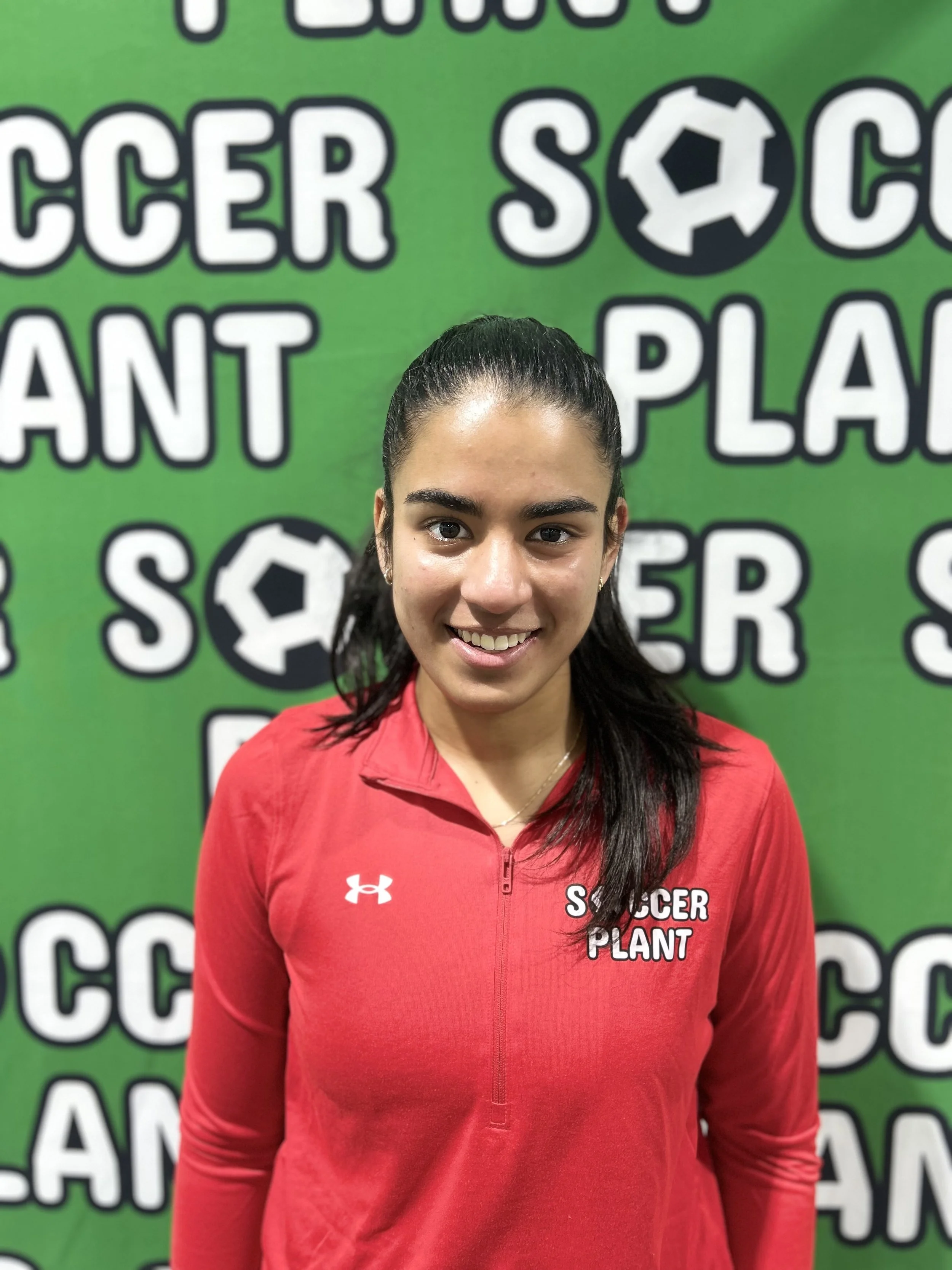 Young woman with dark hair in a ponytail, wearing a red jacket with 'Soccer Plant' logo, standing in front of a green backdrop with white and black text and soccer ball graphics.