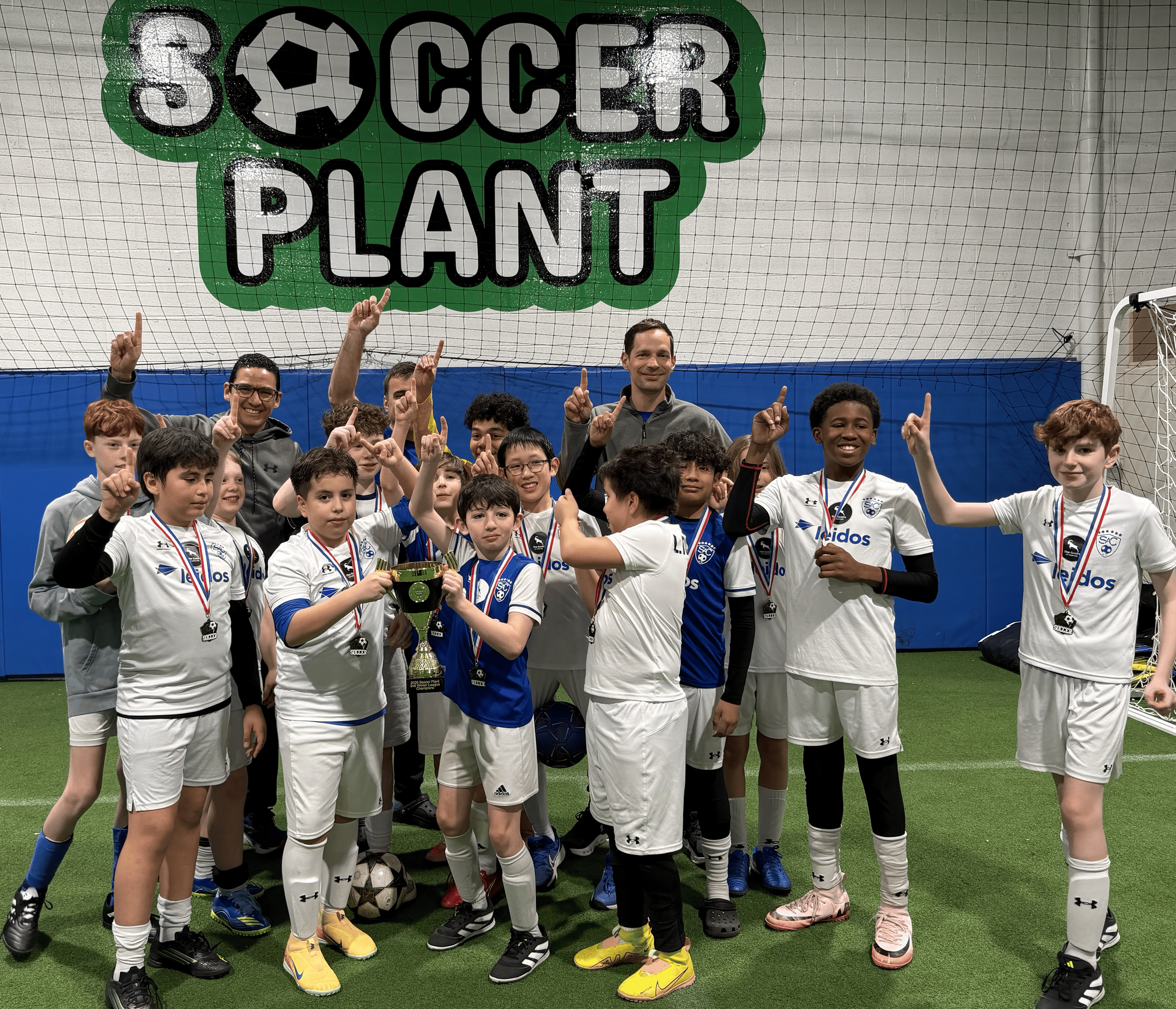 Group of young boys and two adults celebrating a soccer victory inside an indoor soccer facility. They are wearing white soccer uniforms with medals and holding a trophy, standing in front of a sign that reads 'Soccer Plant'.