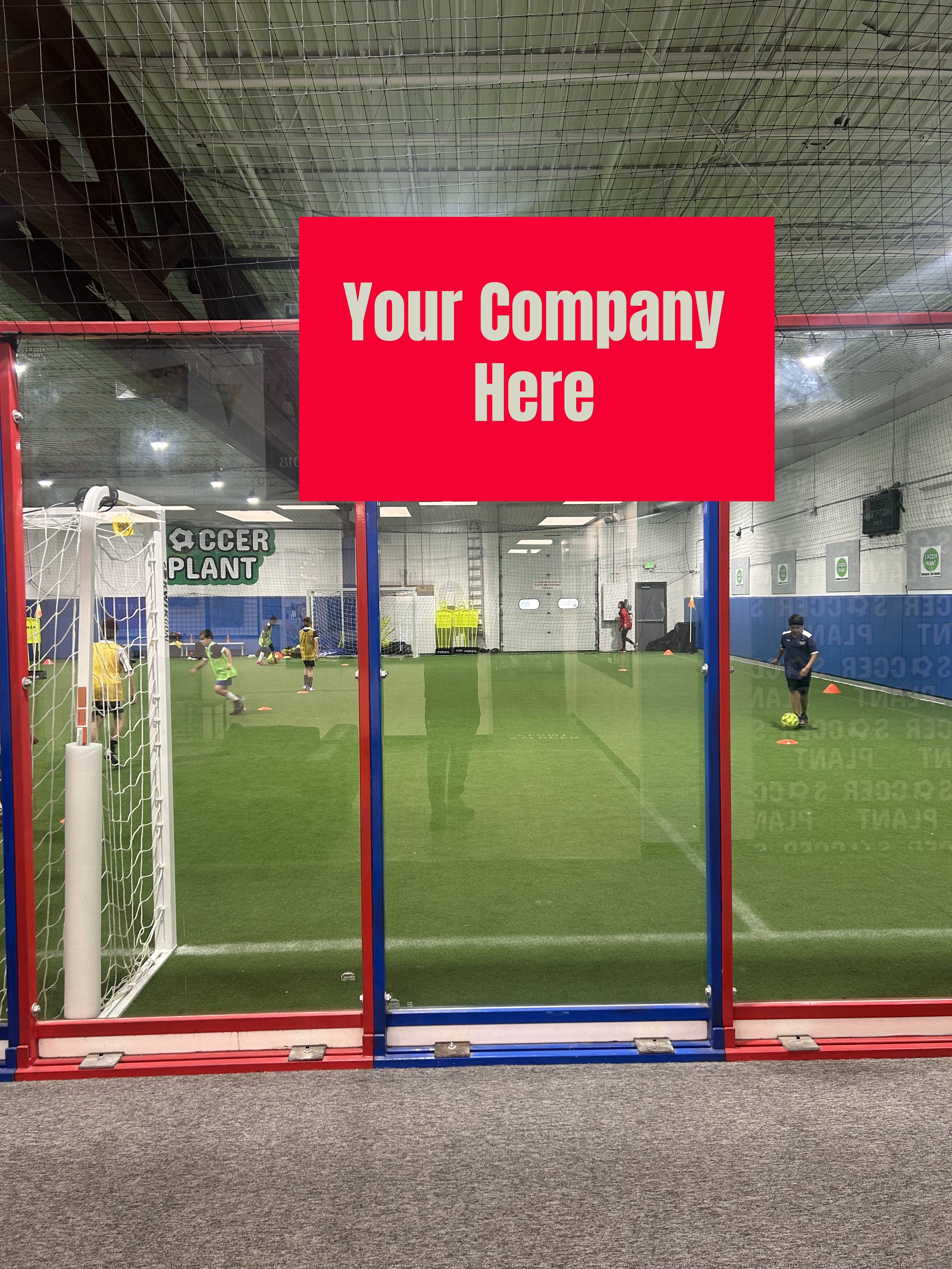 Indoor soccer training facility with children practicing soccer drills behind a glass barrier, with a red sign that reads 'Your Company Here'.