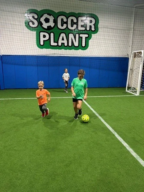 Three children playing soccer inside an indoor facility with a sign that says 'Soccer Plant' above the goal.