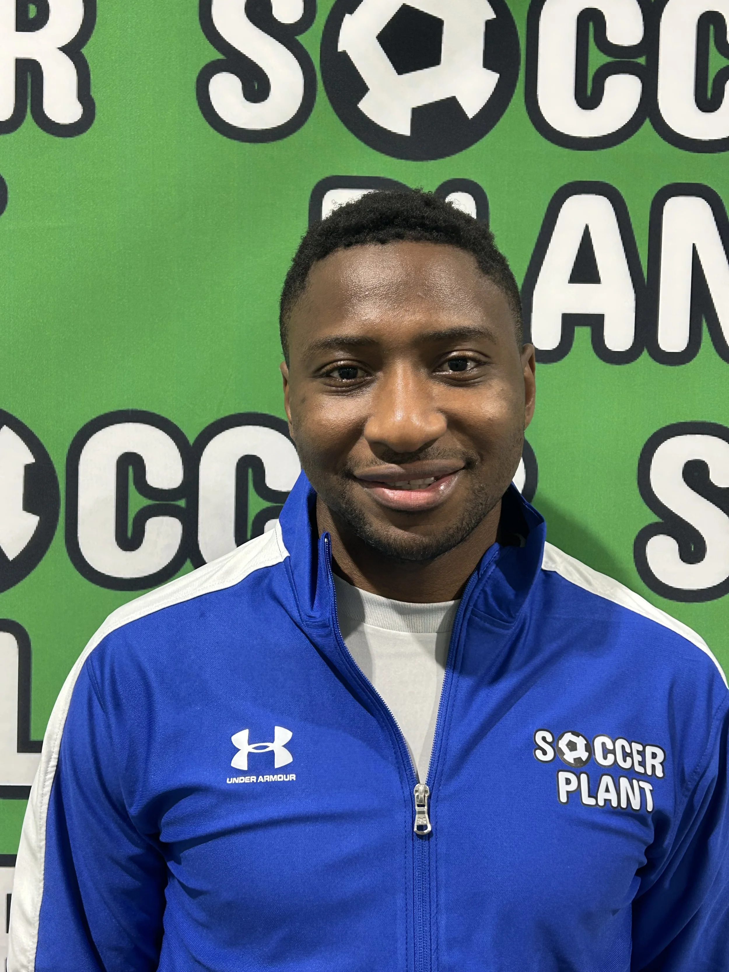 A young man with dark hair and brown skin smiling, wearing a blue Under Armour jacket with 'Soccer Plant' logo on it, standing in front of a green background with large white and black letters and a soccer ball graphic.