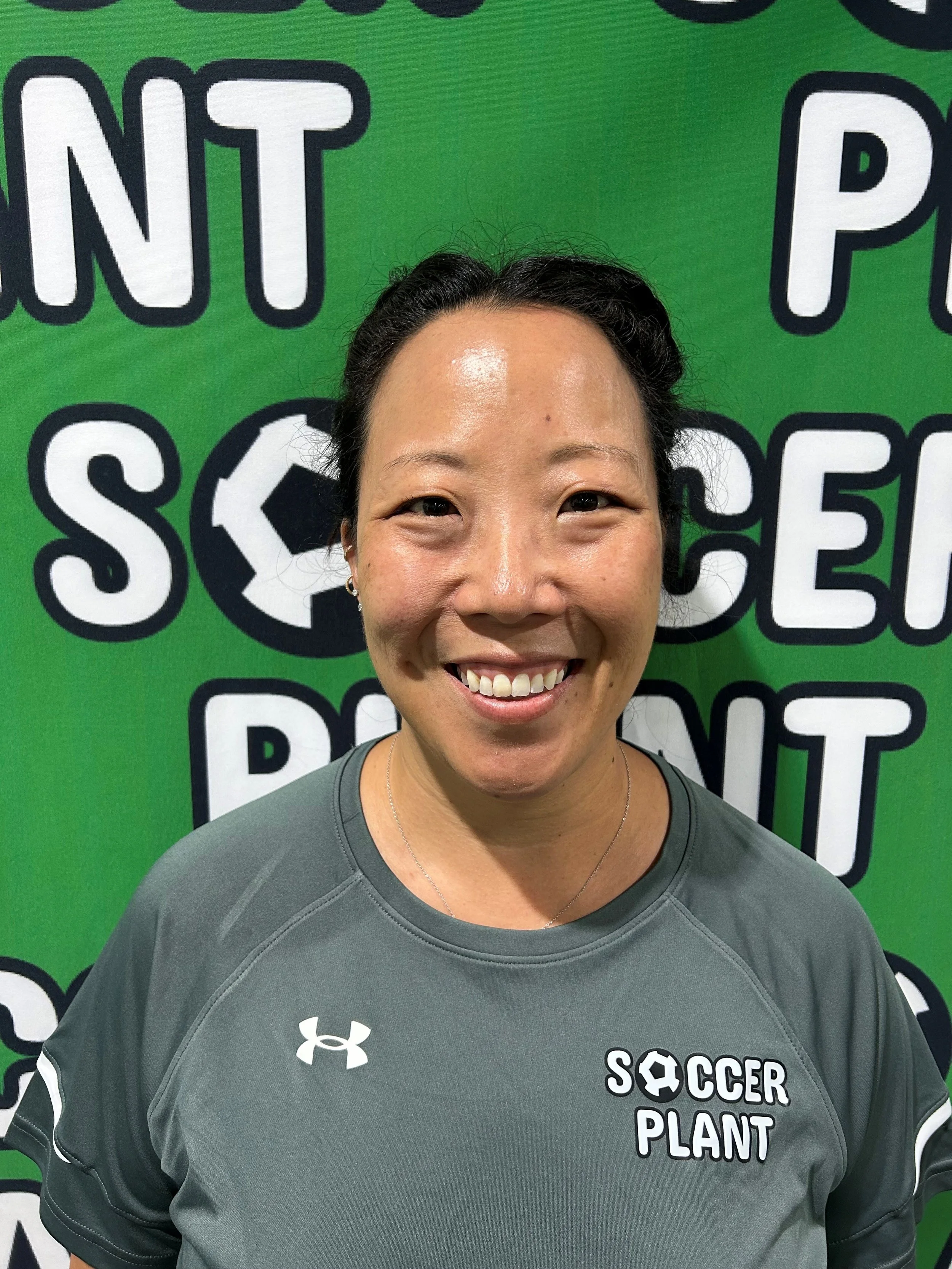 A woman smiling in front of a green background with white and black text that reads "Soccer Planet." She is wearing a gray athletic shirt with the Under Armour logo.
