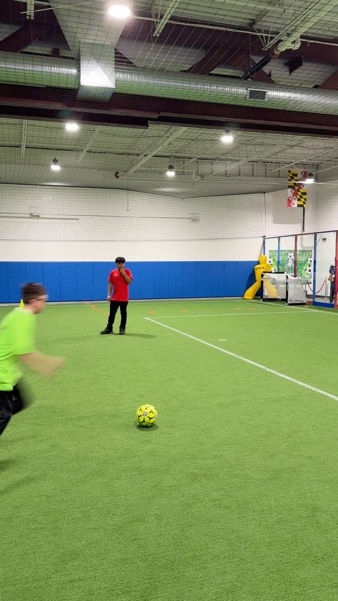 Indoor soccer field with children playing, one child in a neon green shirt kicking a yellow and black soccer ball, another child in a red shirt standing near the field, and a person at the goalpost area near a yellow inflatable structure.