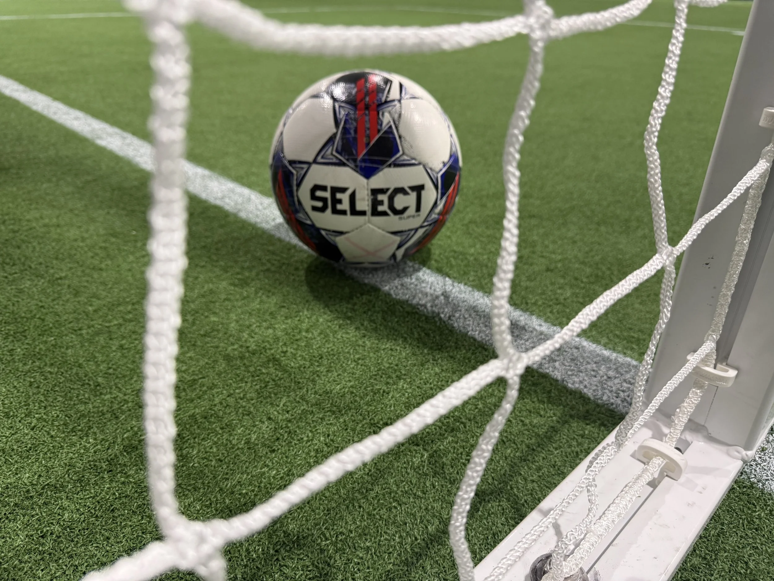 Soccer ball inside the goal on a green artificial turf field with white field lines.