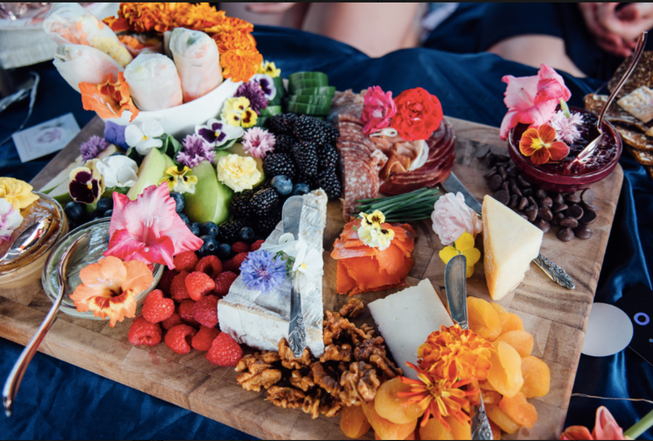 A cheese and fruit platter with edible flowers, various cheeses, fresh berries, nuts, and sliced fruits on a wooden board.