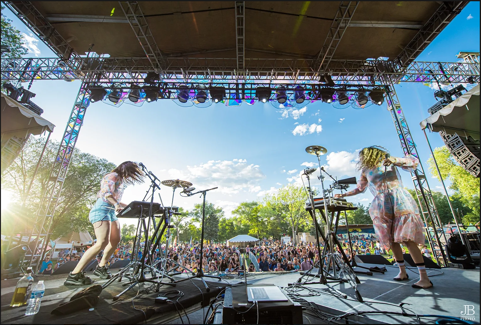 Two female musicians performing on an outdoor stage with a crowd in the background. The stage has lighting and sound equipment, and the performers are surrounded by trees under a blue sky with clouds.