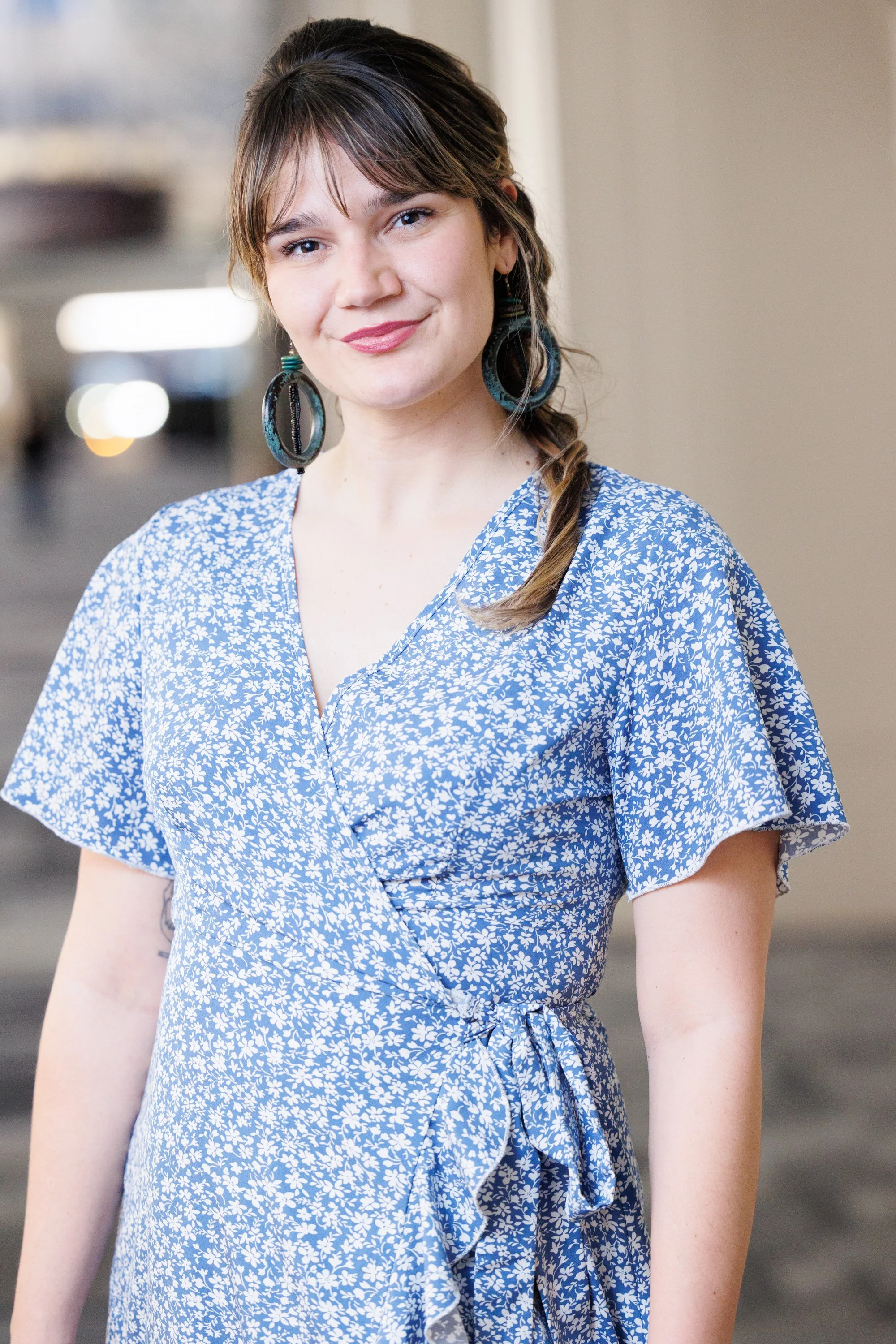 A woman with brown hair styled in a braid, wearing large earrings and a blue and white floral dress, standing outdoors with a blurred background.